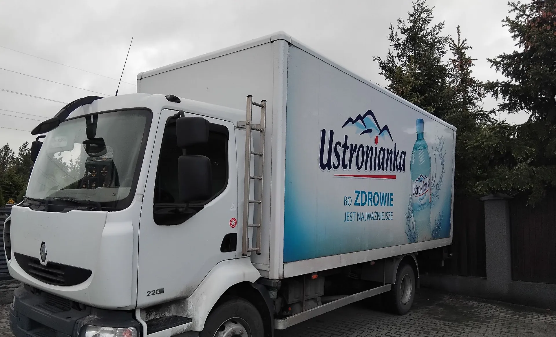 Biała ciężarówka marki Renault z zabudową furgonową. Na boku naczepy znajduje się reklama wody mineralnej Ustronianka z grafiką gór, butelką wody oraz napisem: 'Ustronianka BO ZDROWIE JEST NAJWAŻNIEJSZE'. Pojazd zaparkowany jest na kostce brukowej na tle drzew i szarego nieba.
