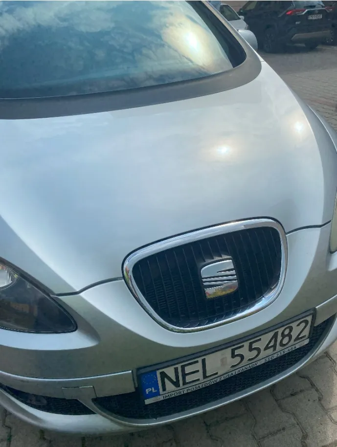 Zbliżenie na przód srebrnego samochodu marki SEAT. Widoczna jest maska, przednia szyba z odbiciem nieba, grill z logo marki oraz tablica rejestracyjna o numerze NEL 55482. Poniżej numeru rejestracyjnego znajduje się napis: 'IMPORT POJAZDÓW Z USA I KANADY 510-235-464'.