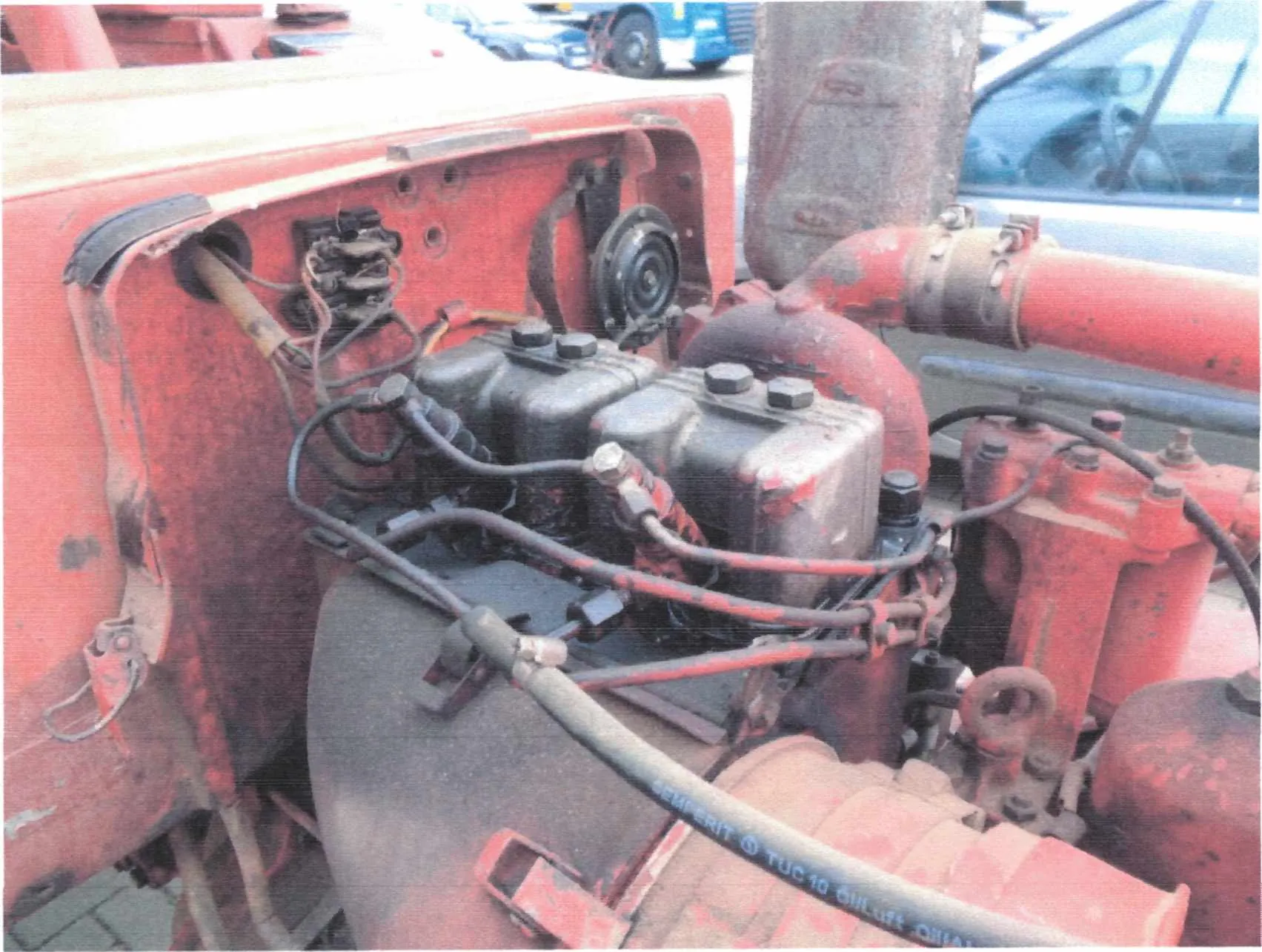 Zbliżenie na czerwony maszynowy agregat z widocznymi czarnymi zbiornikami z nakrętkami, przewodami hydraulicznymi i elektrycznymi, rdzewiejącymi elementami metalowymi oraz dużą czerwoną rurą. W tle rozmyte samochody i pojazdy. Na jednym z przewodów widoczny fragment napisu: 'SEMPERIT TUC 10'.