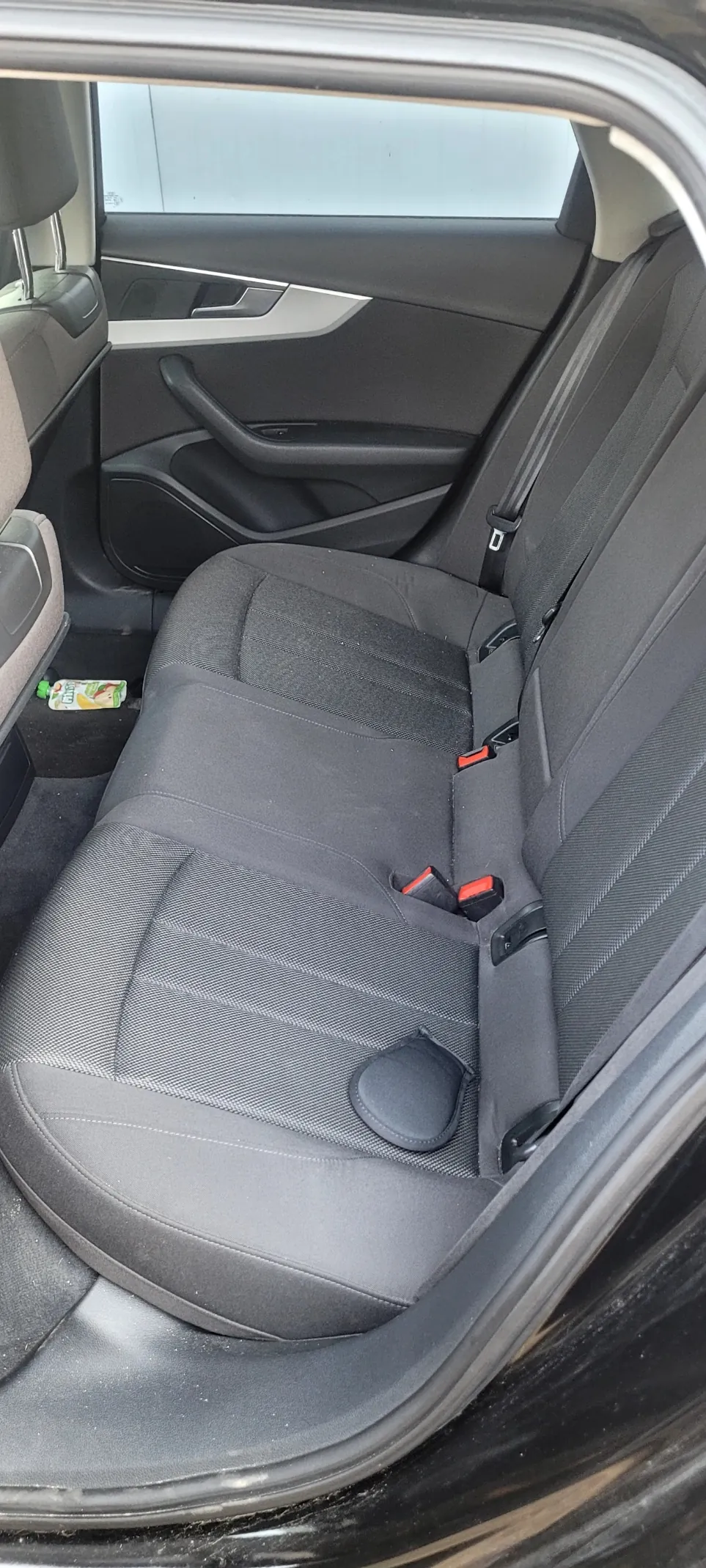 Wnętrze samochodu z widokiem na tylne siedzenie. Szare siedzenia z tkaniny, pasy bezpieczeństwa z czerwonymi przyciskami zwolnienia. Na podłodze po lewej stronie butelka z zielonym korkiem i częściowo widoczną etykietą. Drzwi samochodu z czarnym uchwytem i oknem, przez które widać jasny budynek. Wnętrze w kolorach szarym i czarnym, z widocznymi elementami wykończenia.