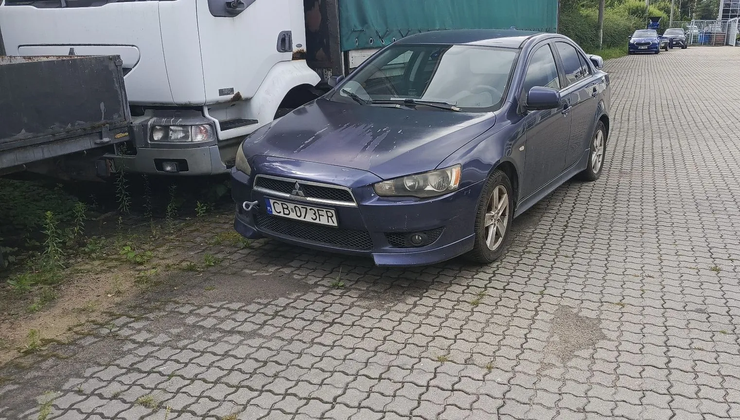 Niebieski samochód osobowy marki Mitsubishi Lancer zaparkowany na kostce brukowej obok białej ciężarówki. Samochód ma polską tablicę rejestracyjną o numerze CB 073FR. Na masce samochodu widoczne są zarysowania i uszkodzenia lakieru.