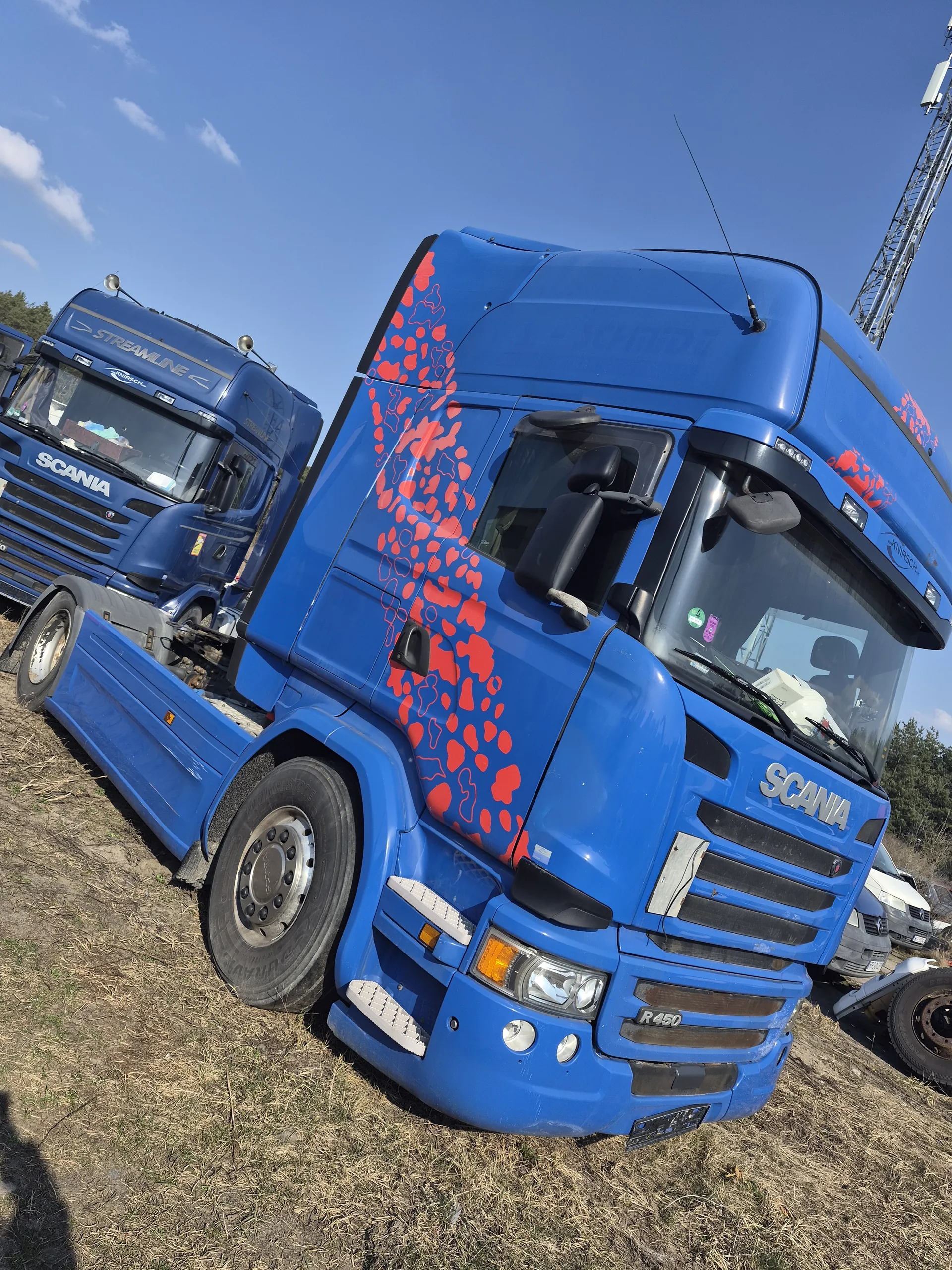 Niebieska ciężarówka marki Scania z czerwonymi wzorami na boku, widoczny napis 'SCANIA' i 'R450' na przodzie. W tle druga niebieska ciężarówka Scania z napisem 'STREAMLINE', sucha trawa, jasne niebo z kilkoma chmurami oraz fragmenty innych pojazdów i żurawia budowlanego.