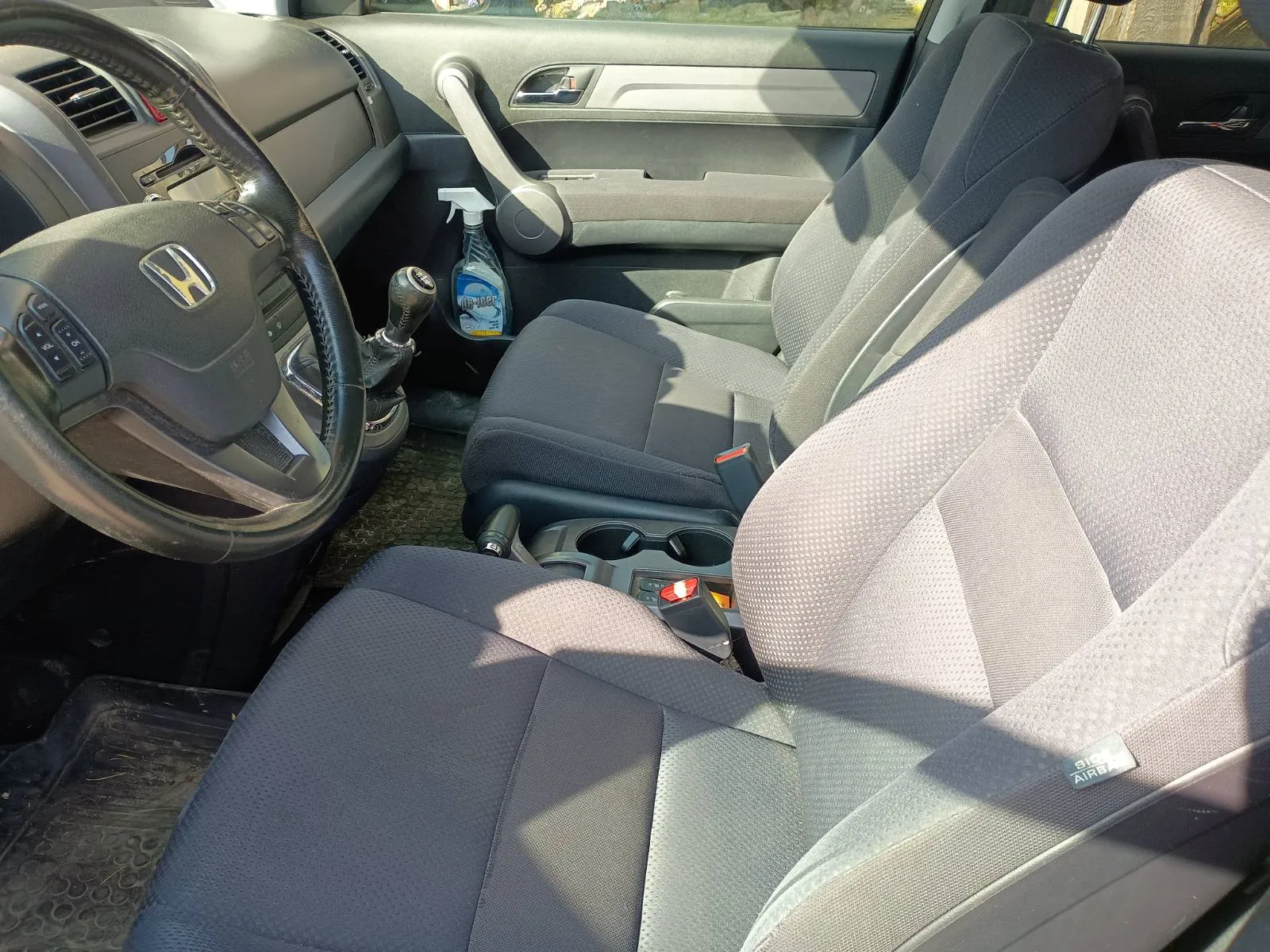 Wnętrze samochodu marki Honda. Widoczna kierownica z logo Hondy, szare siedzenia z drobnym wzorem, dźwignia zmiany biegów, deska rozdzielcza z klimatyzacją oraz butelka sprayu w uchwycie drzwiowym. Na siedzeniu pasażera widoczny napis 'SIDE AIRBAG'. Wnętrze jest jasne, oświetlone światłem słonecznym.