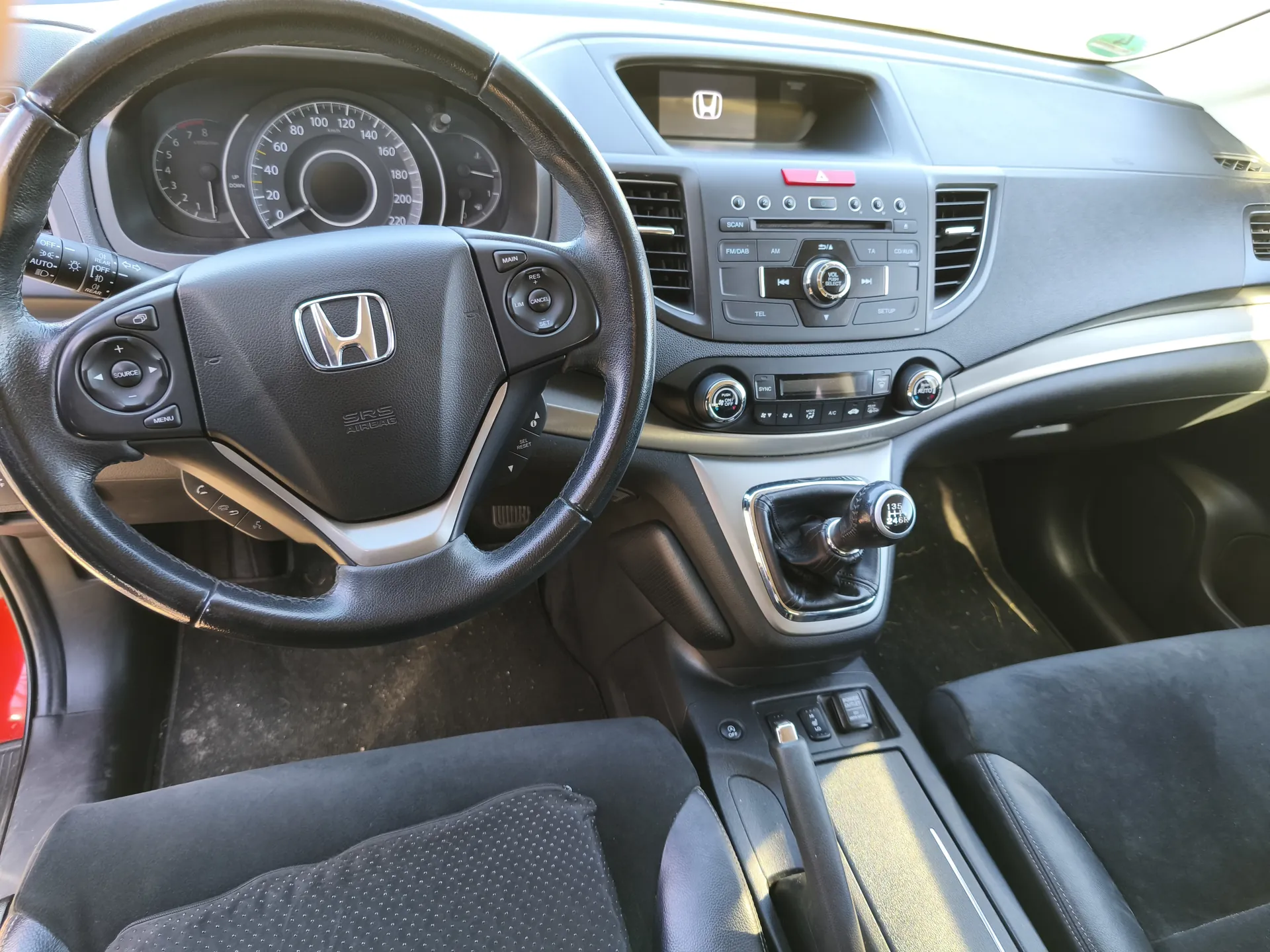 Wnętrze samochodu marki Honda. Widoczna kierownica z logo Hondy i napisem SRS AIRBAG, tablica przyrządów z prędkościomierzem (skala do 220 km/h), centralna konsola z systemem audio, klimatyzacją i przyciskami sterującymi, dźwignia zmiany biegów oraz czarne siedzenia. Wszystkie elementy są prawidłowo ustawione, tekst na przyciskach i wskaźnikach jest czytelny.