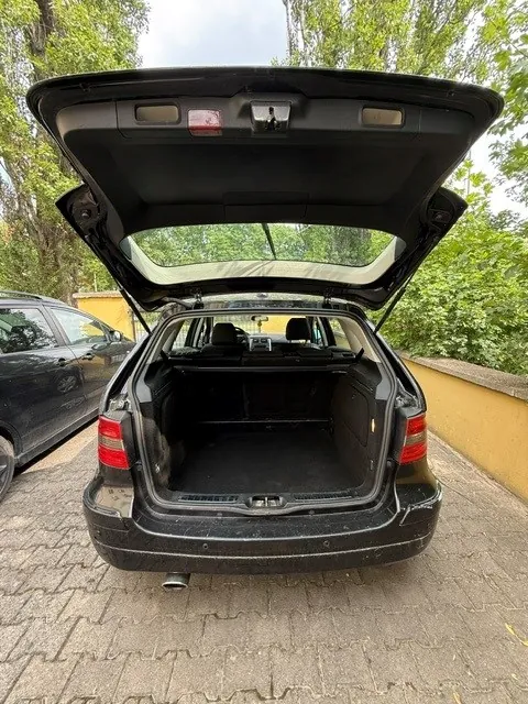 Widok z tyłu czarnego samochodu typu kombi z otwartą klapą bagażnika. Wnętrze bagażnika jest puste i wyłożone czarnym materiałem. Samochód zaparkowany jest na kostce brukowej obok żółtego murka i zielonych drzew. Po lewej stronie widoczny jest fragment innego, ciemnego samochodu.