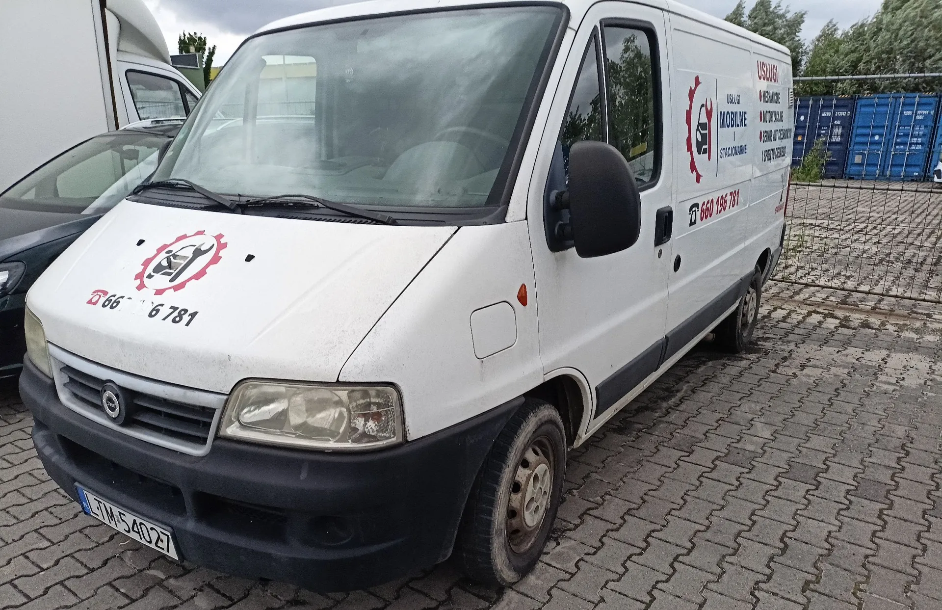 Biały furgon marki Opel zaparkowany na kostce brukowej. Na masce samochodu znajduje się czerwone logo z kluczem francuskim i numerem telefonu 660 196 781. Na boku pojazdu widnieje napis 'USŁUGI MOBILNE' wraz z listą usług i numerem telefonu 660 196 781. Samochód ma tablicę rejestracyjną o numerze LTM 54027.