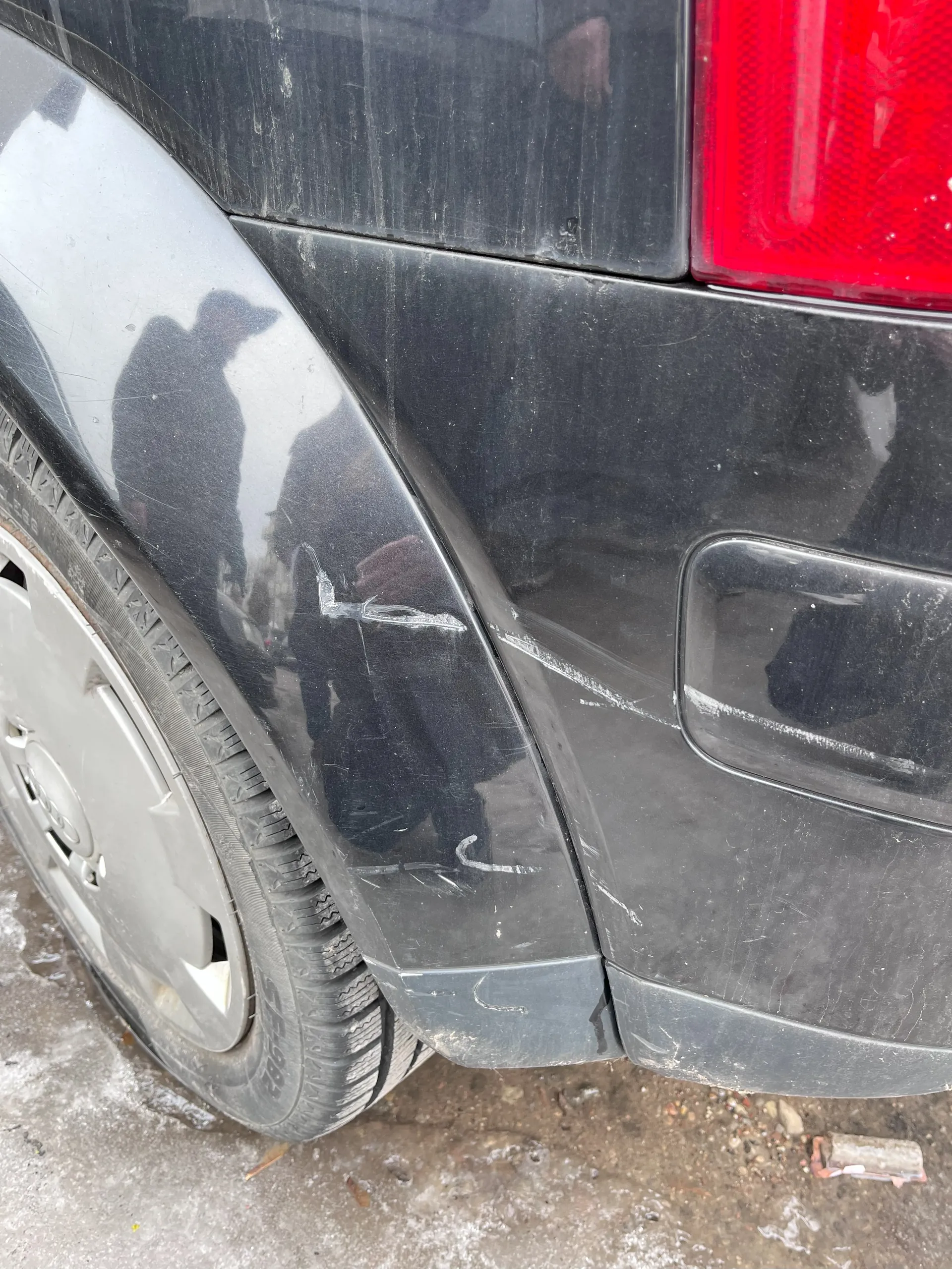 Zbliżenie na tylną część czarnego samochodu. Widoczne są zadrapania na błotniku i nadkolu, czerwone światło tylnego reflektora oraz srebrna felga z oponą. Na powierzchni nadkola odbijają się sylwetki ludzi. Pod samochodem znajduje się ziemia z resztkami śniegu lub lodu.