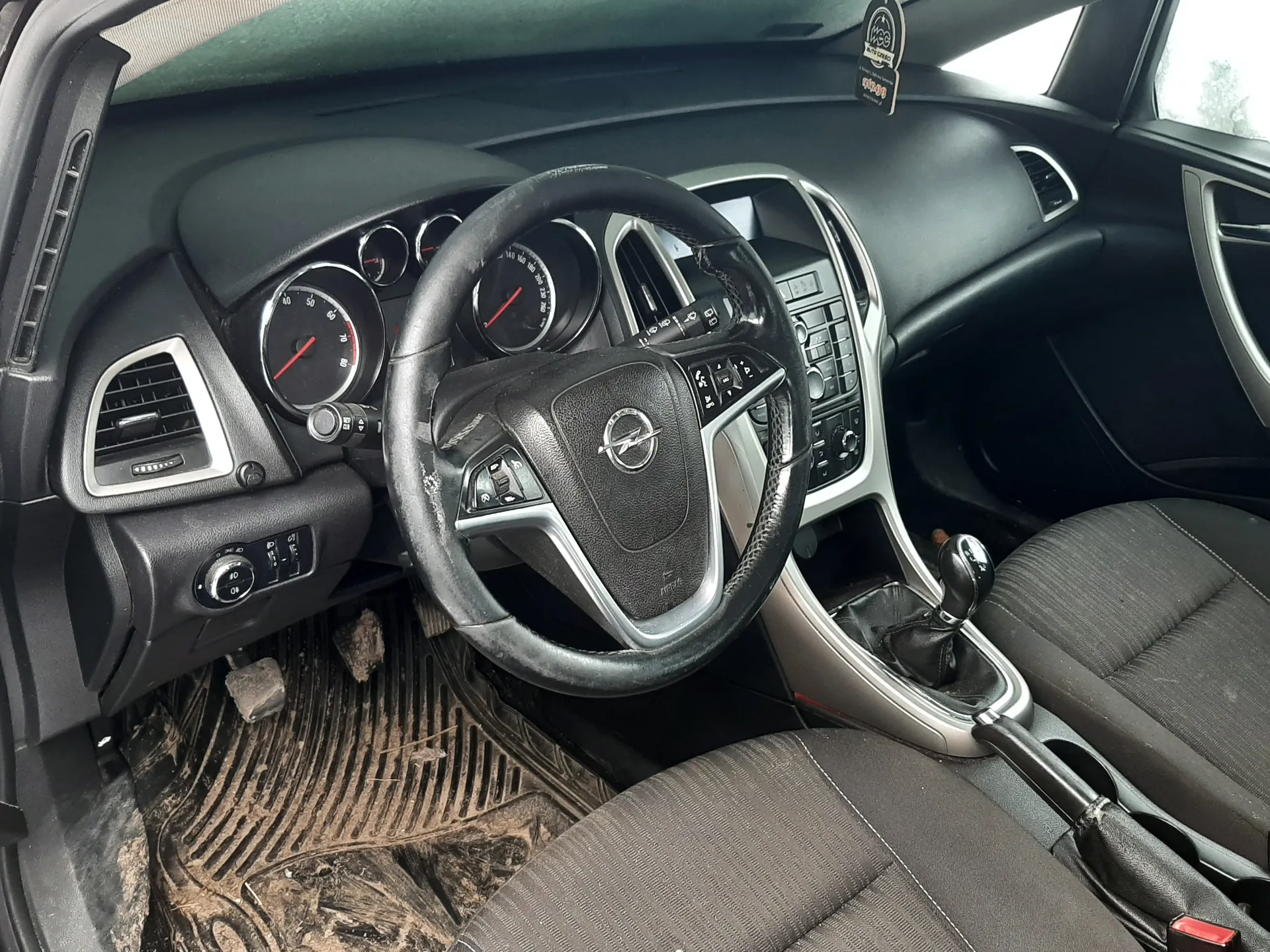 Wnętrze samochodu Opel z czarną kierownicą z logo, analogowymi wskaźnikami prędkości i obrotomierza, szarymi siedzeniami. Brudne dywany podłogowe, przyciski na konsoli środkowej, boczne wywietrzniki. Na szybie naklejka z napisem "MOT 4990".