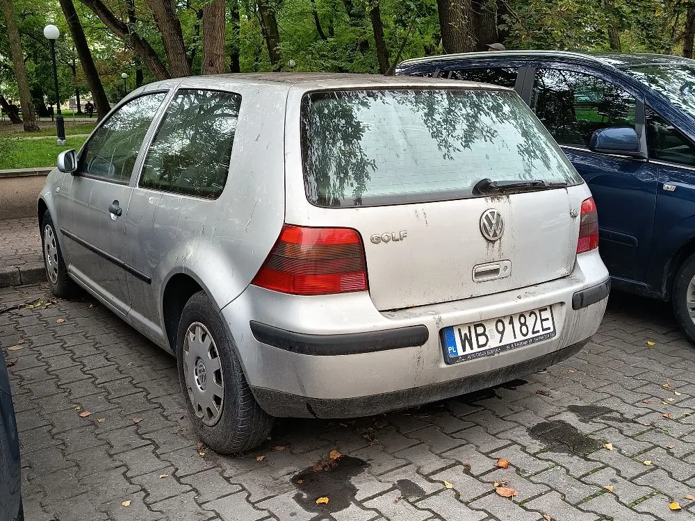 Srebrny samochód marki Volkswagen Golf zaparkowany tyłem na kostce brukowej w otoczeniu zieleni i drzew. Samochód jest zabrudzony, szczególnie w dolnej części nadwozia i na klapie bagażnika. Widoczny jest napis GOLF oraz logo Volkswagena. Tablica rejestracyjna samochodu to WB 9182E. Obok stoi ciemnoniebieski samochód.