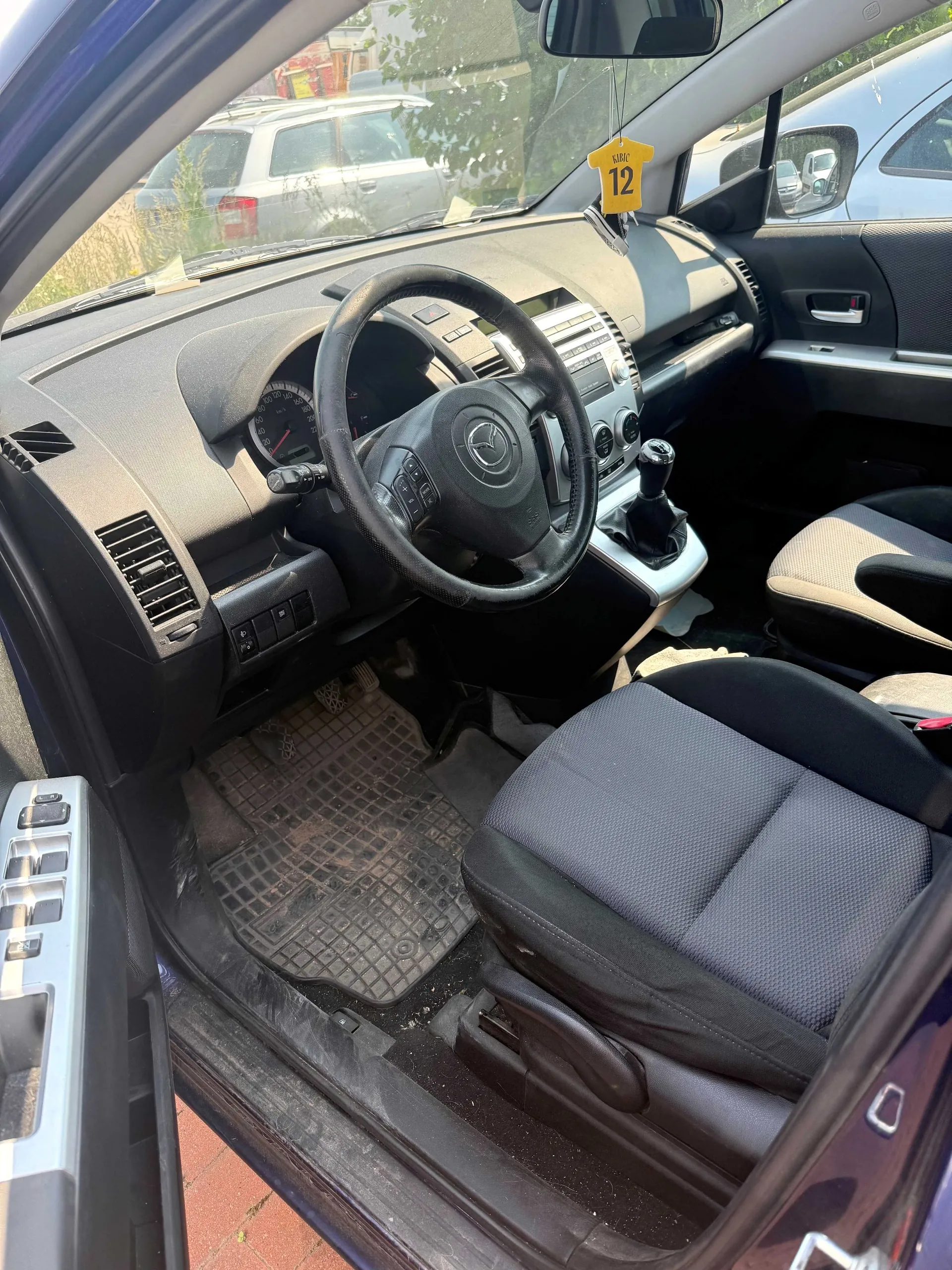Wnętrze samochodu marki Mazda widziane z perspektywy otwartych drzwi kierowcy. Widoczna jest czarna kierownica z logo Mazdy, deska rozdzielcza, lewarek zmiany biegów oraz ciemnoszare fotele materiałowe. Na podłodze znajduje się gumowa mata z wypustkami. Przez przednią szybę widoczny jest fragment innego, srebrnego samochodu oraz żółta zawieszka z numerem 12.