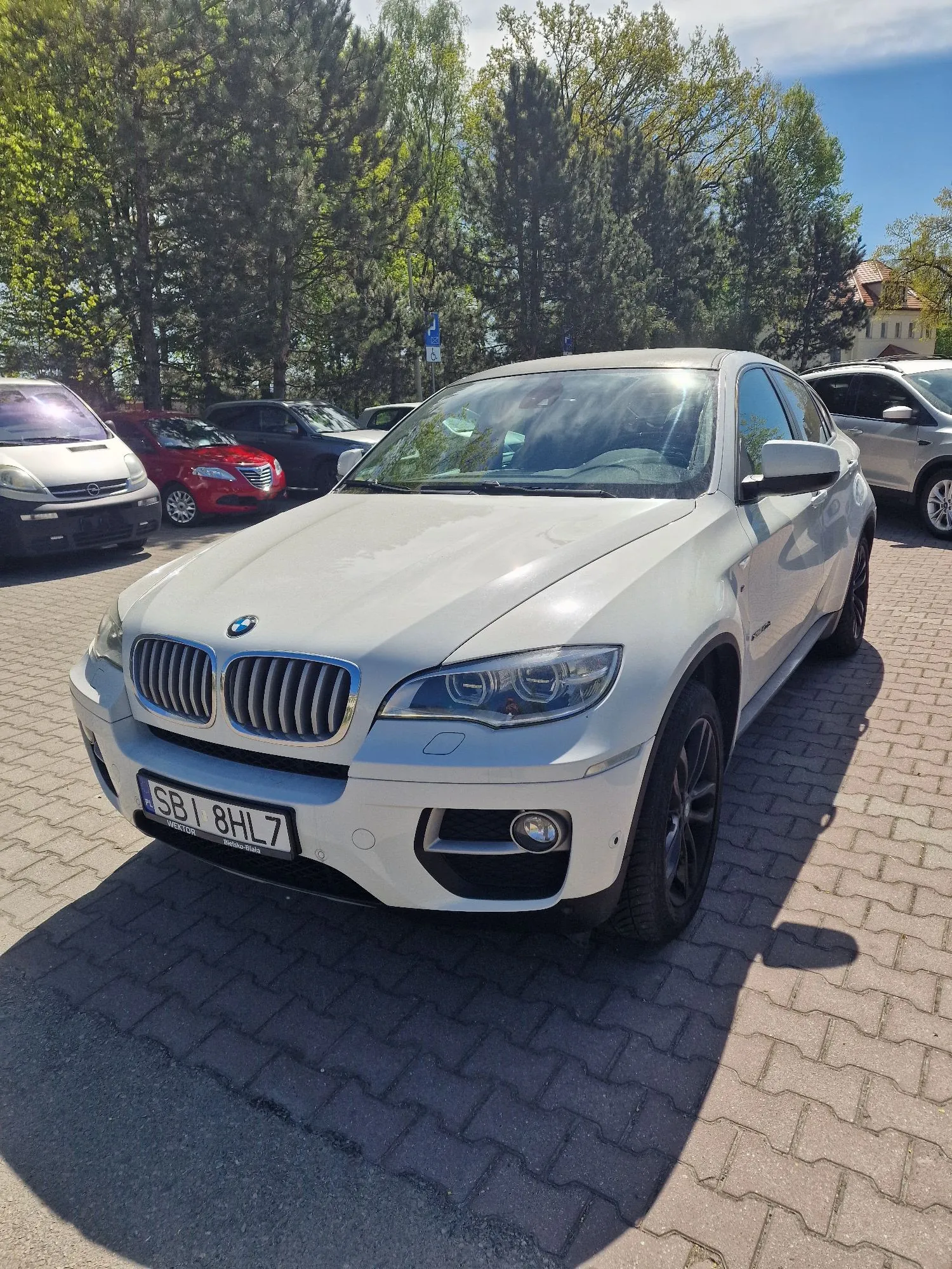 Biały samochód marki BMW zaparkowany na kostce brukowej. Pojazd jest widoczny z przodu pod kątem. Na samochodzie znajduje się tablica rejestracyjna o treści SBI 8HL7. W tle widoczne są inne zaparkowane samochody w kolorach srebrnym, czerwonym i ciemnoszarym oraz zielone drzewa pod niebieskim niebem.