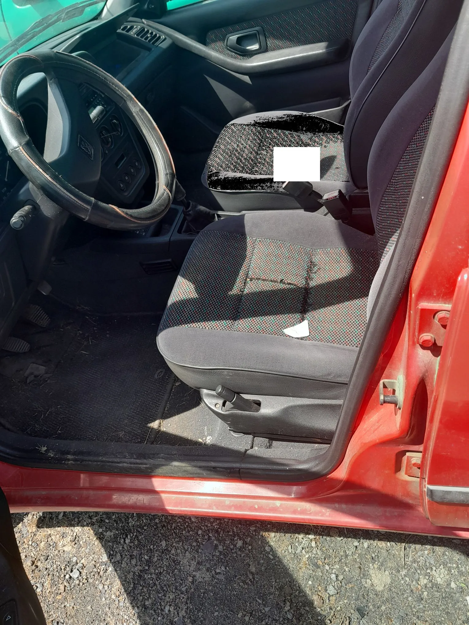 Wnętrze samochodu z perspektywy otwartych drzwi pasażera. Widoczny jest czarny fotel z materiałem w drobną, czerwoną kratkę. Siedzisko fotela jest przetarte i uszkodzone w centralnej części. W tle widać czarną kierownicę, deskę rozdzielczą oraz fragment czerwonego nadwozia samochodu.