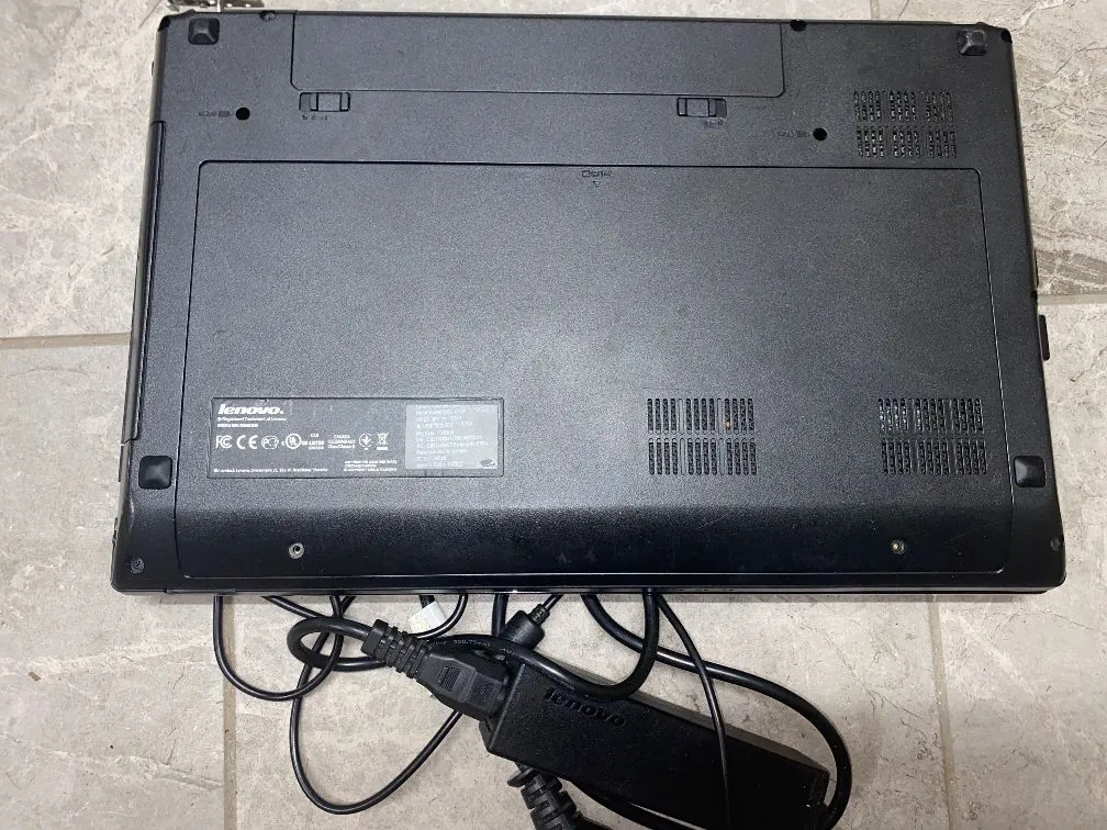 Widok od spodu czarnego laptopa marki Lenovo, leżącego na jasnej, kafelkowej powierzchni. Na obudowie widoczne są otwory wentylacyjne, gumowe stopki oraz tabliczka znamionowa z logo Lenovo i certyfikatami. Pod laptopem znajduje się czarny zasilacz z kablem i logo Lenovo.