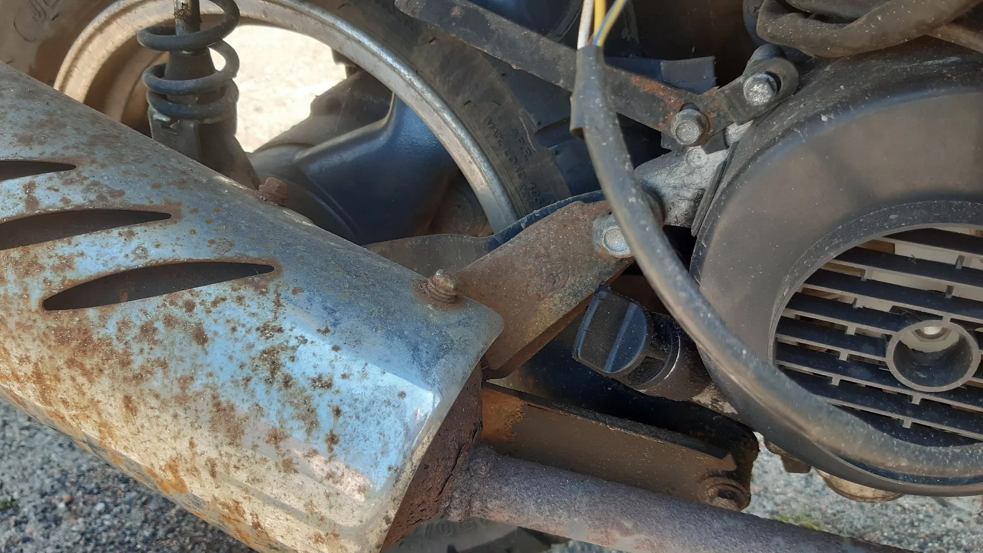 Zbliżenie na tylną część motocykla lub skutera. Widoczny jest zardzewiały, metalowy tłumik z otworami wentylacyjnymi, fragment koła z oponą, amortyzator sprężynowy oraz czarna obudowa silnika z kratką wentylacyjną.