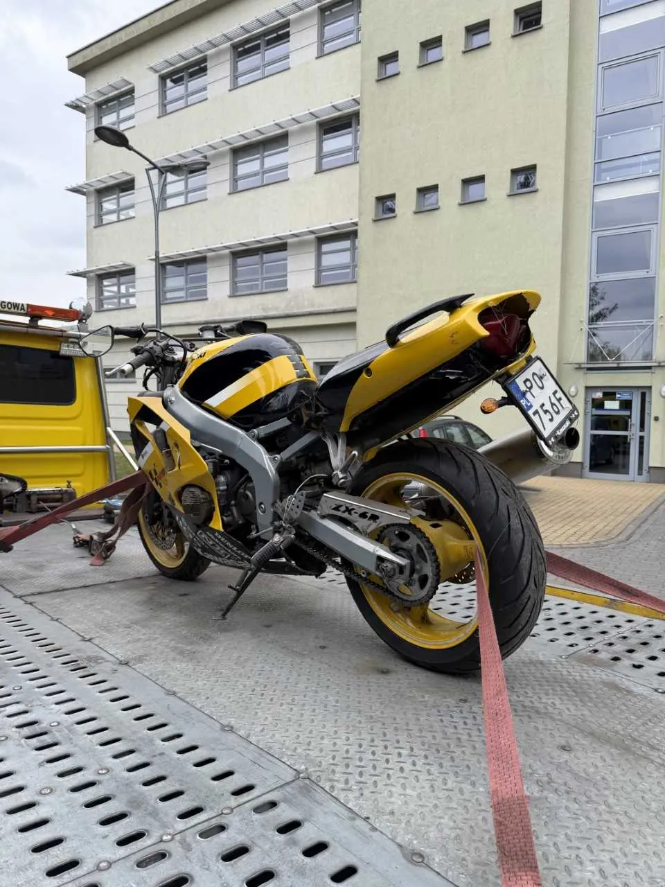 Zdjęcie przedstawia żółto-czarny motocykl sportowy z tablicą rejestracyjną PO 756F, stojący na metalowej platformie z perforacjami. Motocykl jest przywiązany czerwonymi paskami do żółtego pojazdu z napisem GOWA widocznym na boku. W tle znajduje się wielopiętrowy budynek o beżowej elewacji z oknami, latarnia uliczna oraz fragment chodnika. Na motocyklu widoczne są elementy takie jak wydech, koła z żółtymi felgami oraz napis ZX-6R na ramie.