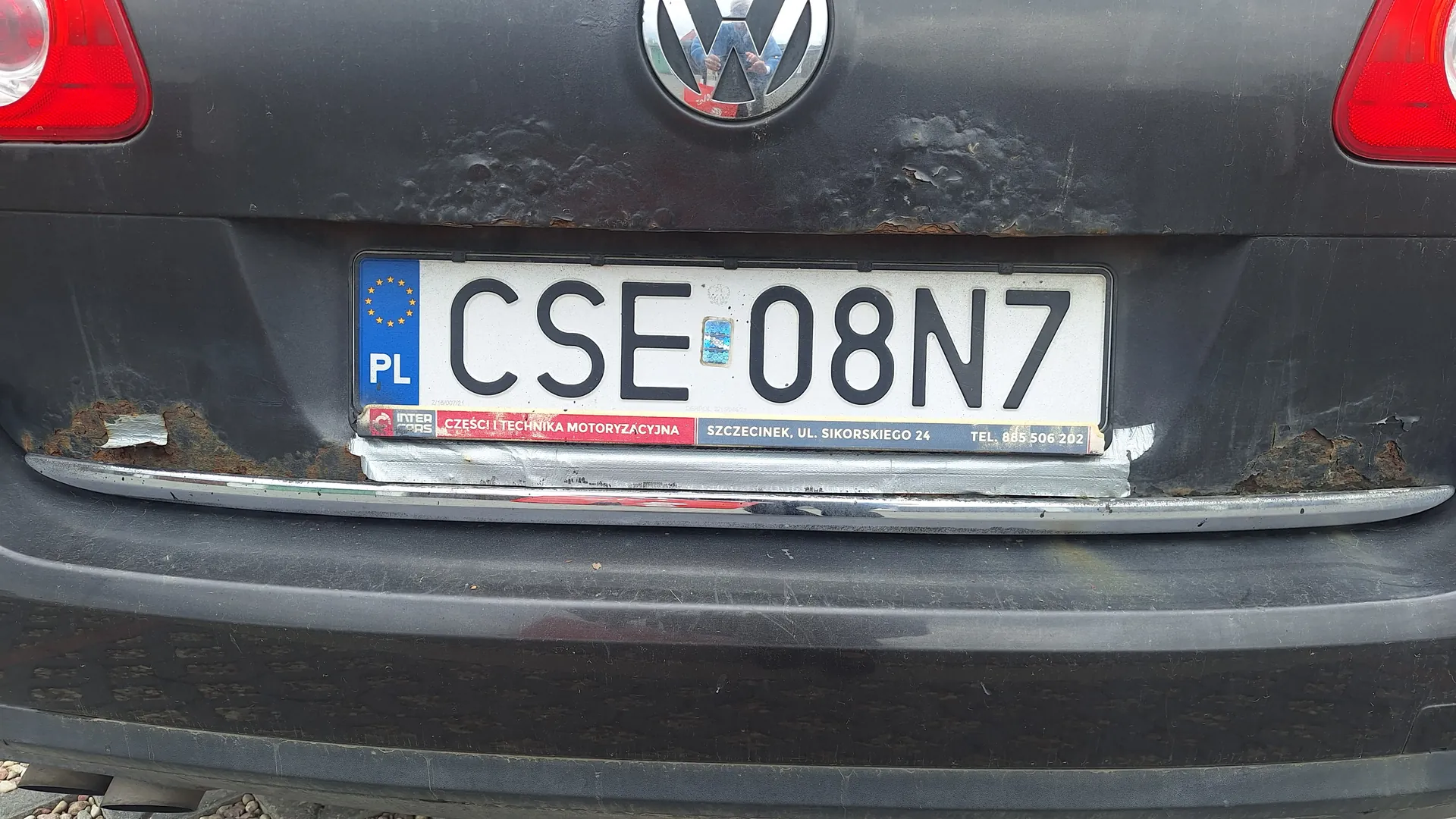 Tył ciemnego samochodu z tablicą rejestracyjną oznaczoną CSE 08N7. Tablica posiada niebieski pas z napisem PL i flagą Unii Europejskiej po prawej stronie. Widoczny jest logo Volkswagena oraz czerwone światła tylnego. Wokół tablicy widoczne są ślady rdzy. Na górnej krawędzi tablicy znajdują się naklejki z tekstem, w tym częściowo czytelnym: SZCZECINIEK, UL. SIKORSKIEGO 24.