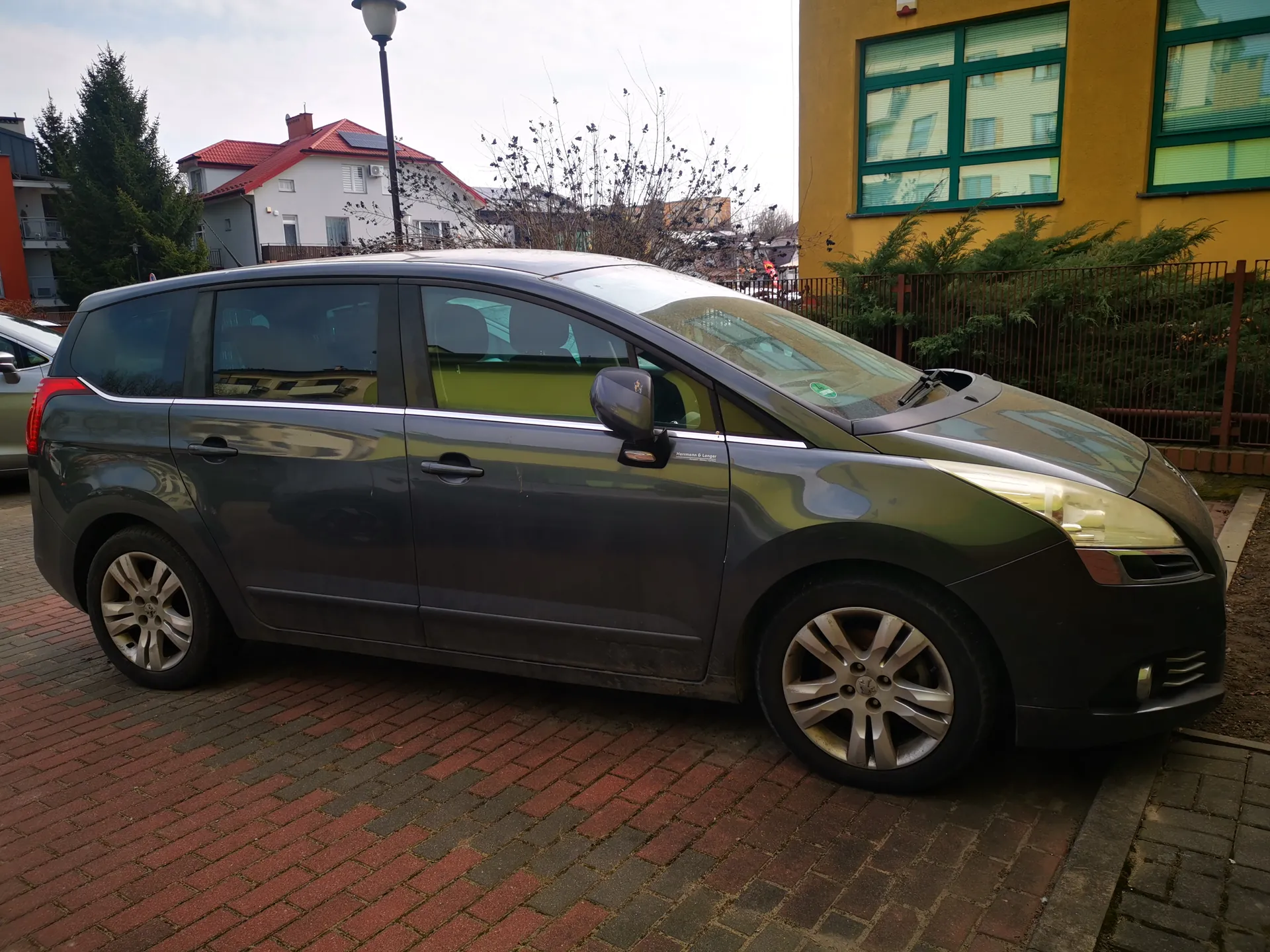 Ciemnoszary minivan zaparkowany na czerwono-szarym brukowanym placu. W tle żółty budynek z zielonymi ramami okiennymi, białe domy z czerwonymi dachami, latarnia uliczna, drzewa i ogrodzenie. Niebo jest pochmurne.