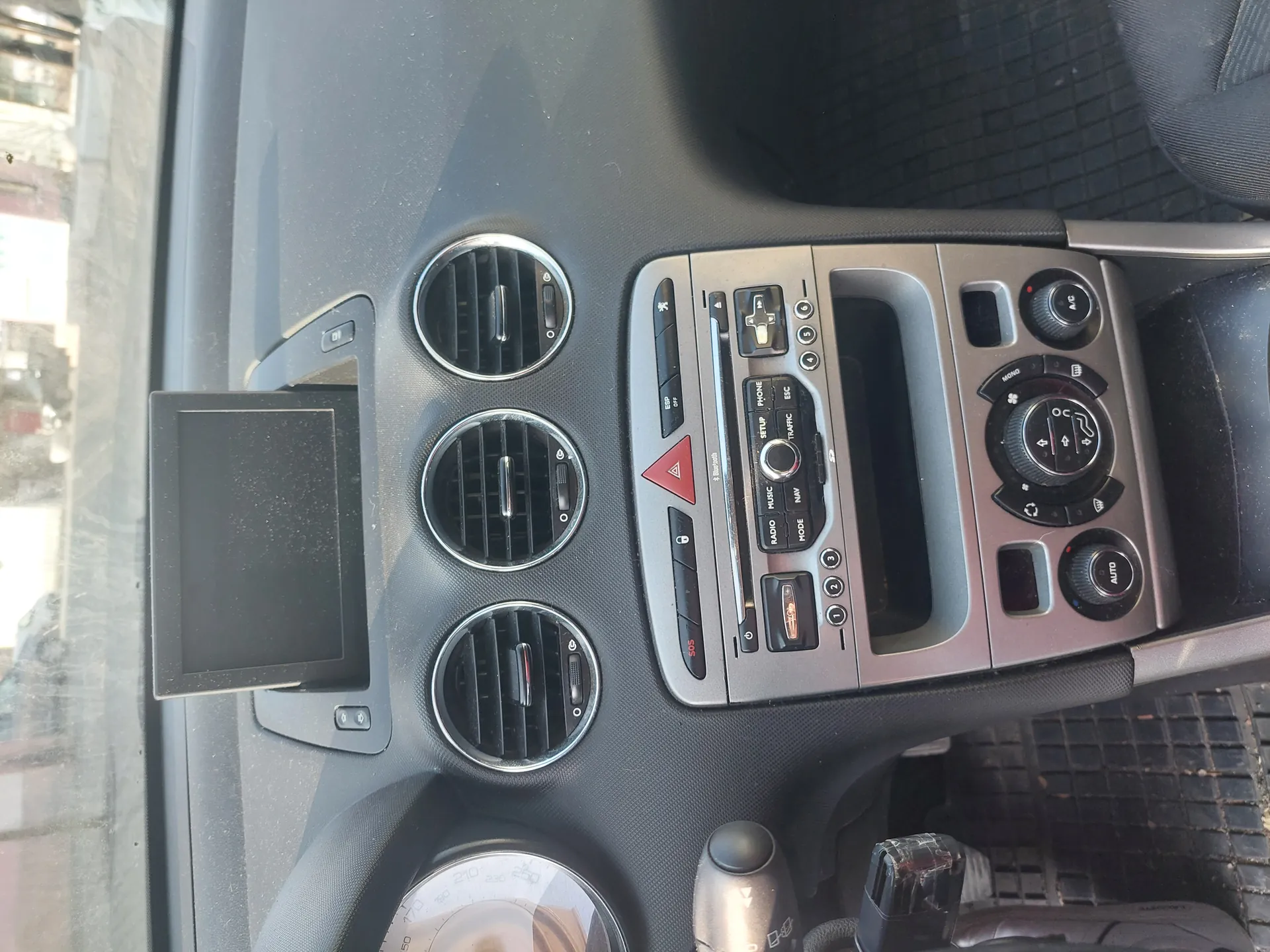 Wnętrze samochodu z widokiem na deskę rozdzielczą. Trzy okrągłe wywiewacze powietrza po lewej stronie, mały czarny ekran obok nich. W centrum konsoli środkowej przyciski sterujące, w tym czerwony trójkątny przycisk awaryjny, przyciski oznaczone 'RADIO', 'MODE', 'AUTO' oraz pokrętła klimatyzacji. Widoczna również dźwignia zmiany biegów i fragment wskaźnika prędkości. W tle przez przednią szybę widoczne budynki.