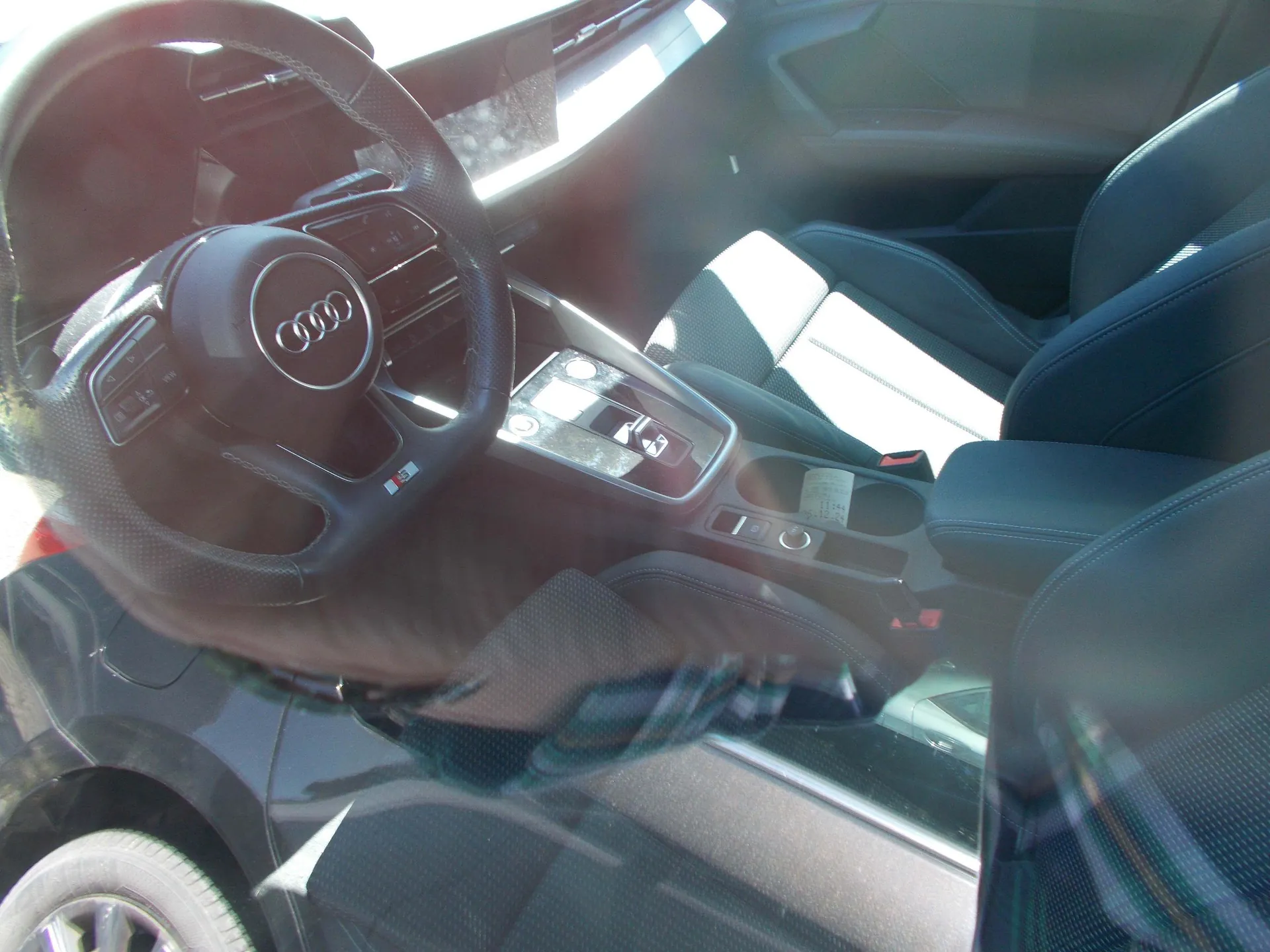 Zdjęcie wnętrza samochodu marki Audi, wykonane przez szybę. Widoczna jest kierownica z logo Audi, konsola środkowa z lewarkiem zmiany biegów oraz przednie fotele z ciemnej tapicerki. W dolnej części zdjęcia widoczny jest fragment przedniego koła i nadkola samochodu.