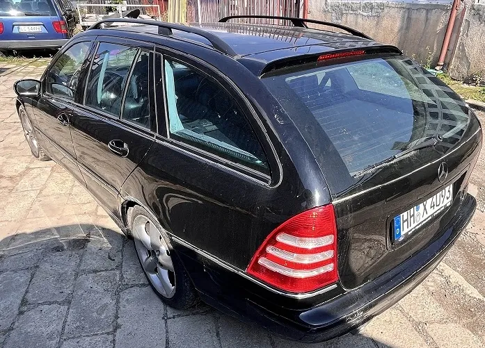 Czarny samochód marki Mercedes-Benz w nadwoziu kombi, zaparkowany na kostce brukowej. Widok z tyłu pod kątem. Samochód posiada relingi dachowe oraz niemiecką tablicę rejestracyjną o numerze HH X 4093. W tle widoczny jest fragment innego niebieskiego samochodu oraz szary mur.