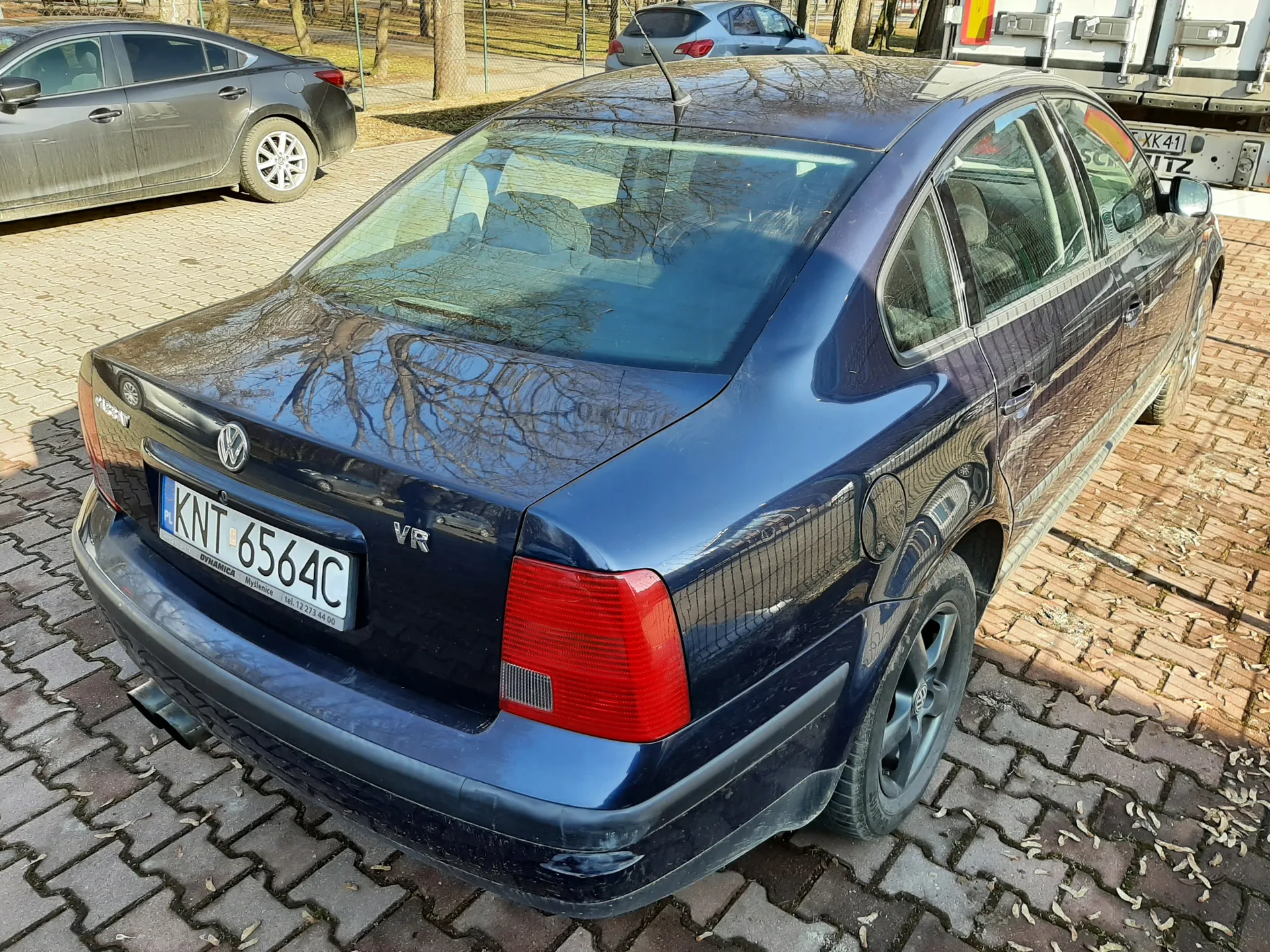 Ciemnoniebieski samochód osobowy marki Volkswagen Passat z widocznym tyłem, tablicą rejestracyjną PL KNT 6564C oraz napisem VR na bagażniku. W tle inne pojazdy: szary sedan po lewej, srebrny samochód i ciężarówka z tablicą XK 41 TIZ po prawej. Samochód zaparkowany na brukowanej powierzchni z rozsypanymi liśćmi, w tle drzewa i ogrodzenie. Na tablicy rejestracyjnej widoczny napis DYNAMIC oraz numer telefonu tel. 12 273 44 00.