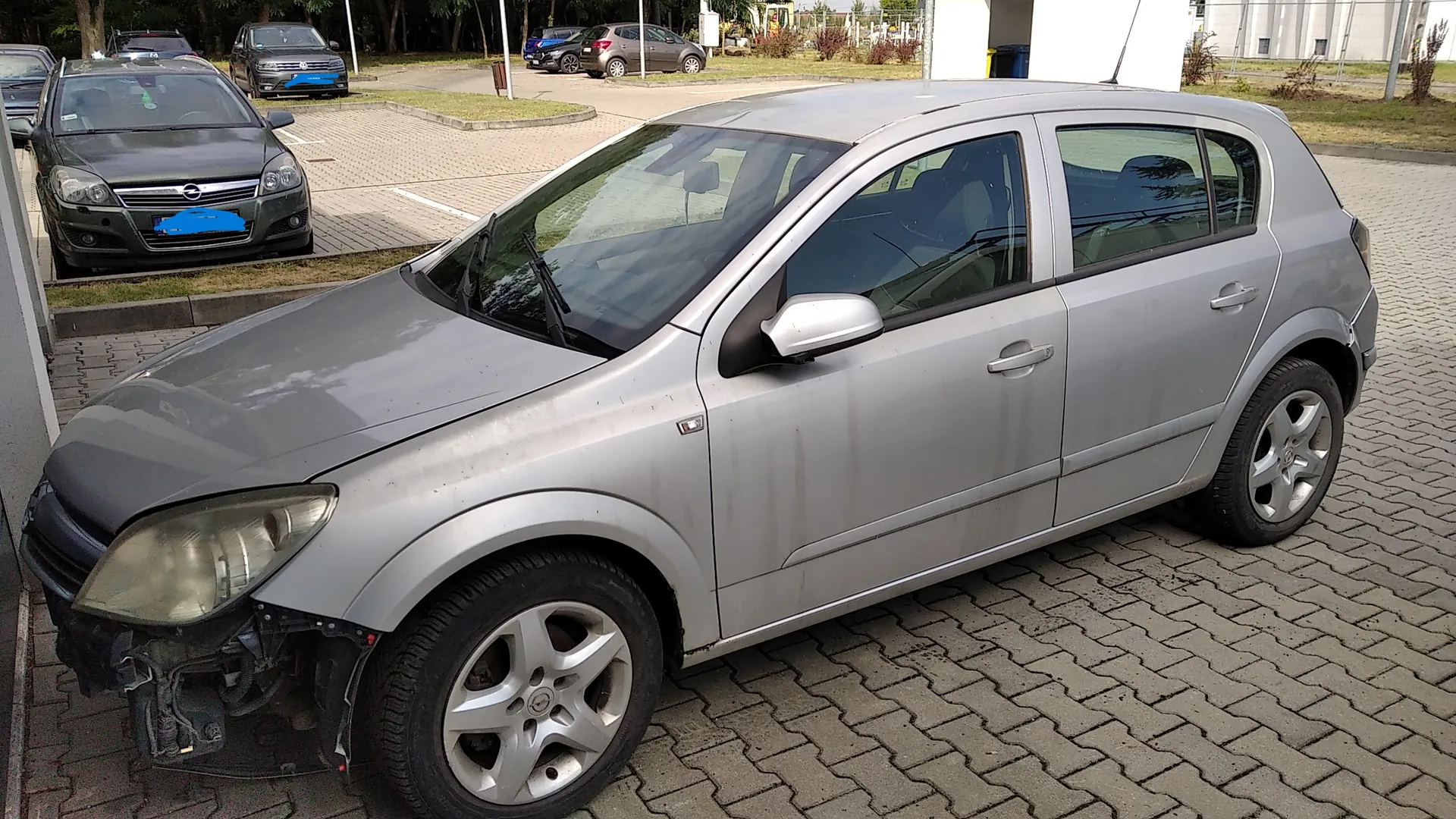 Srebrny samochód osobowy marki Opel Astra w nadwoziu hatchback, zaparkowany na kostce brukowej. Pojazd ma uszkodzony przedni zderzak po lewej stronie, który jest częściowo oderwany, odsłaniając elementy konstrukcyjne. W tle widoczne są inne zaparkowane samochody oraz pas zieleni z drzewami.