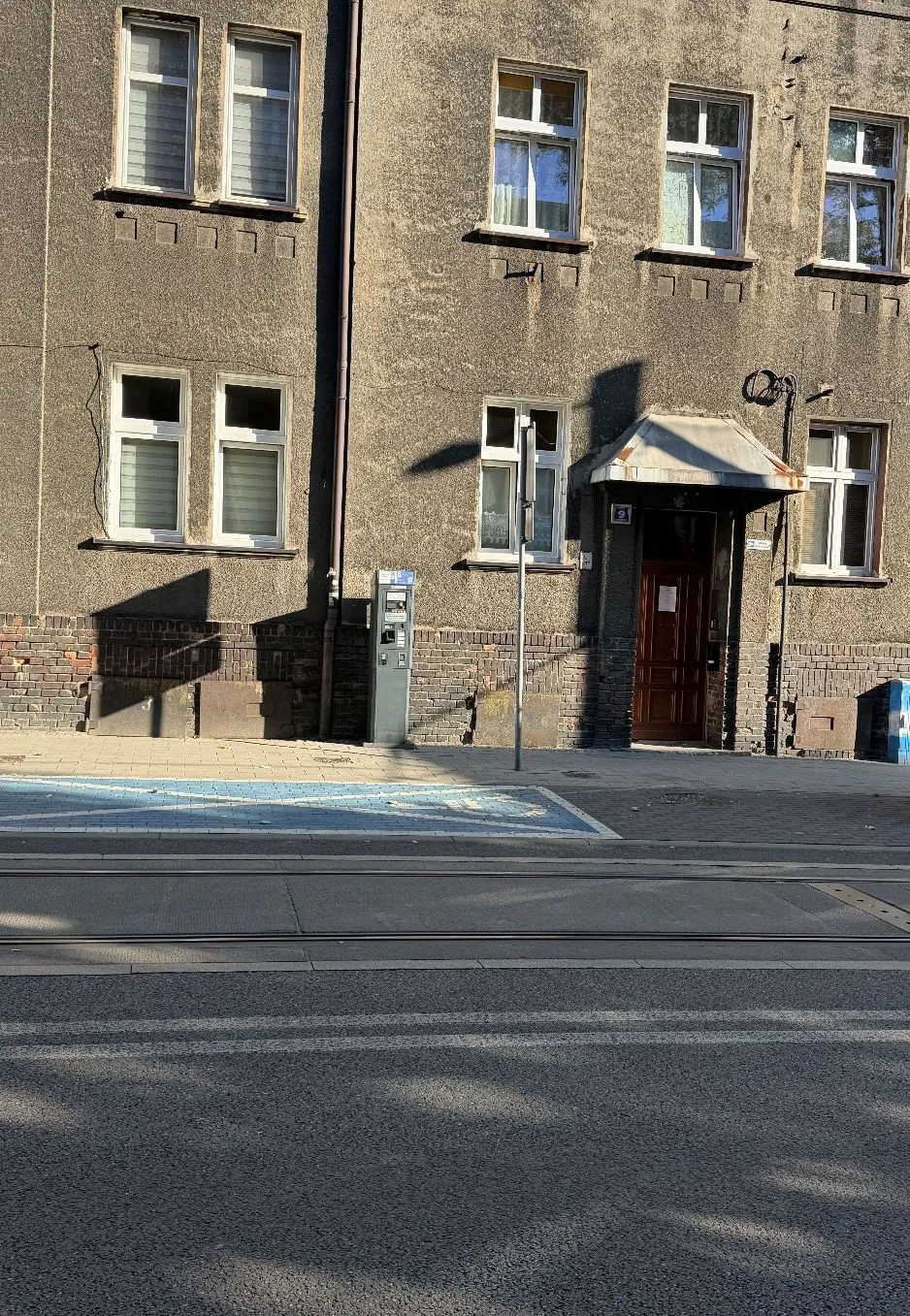 Elewacja szarego, tynkowanego budynku z kilkoma oknami w dwóch rzędach. Na parterze znajduje się wejście z ciemnobrązowymi drzwiami, nad którymi jest mały daszek. Obok drzwi stoi szary automat parkingowy. Przed budynkiem widoczny jest chodnik, niebieski obszar wyznaczający miejsce parkingowe oraz fragment ulicy z szynami tramwajowymi.