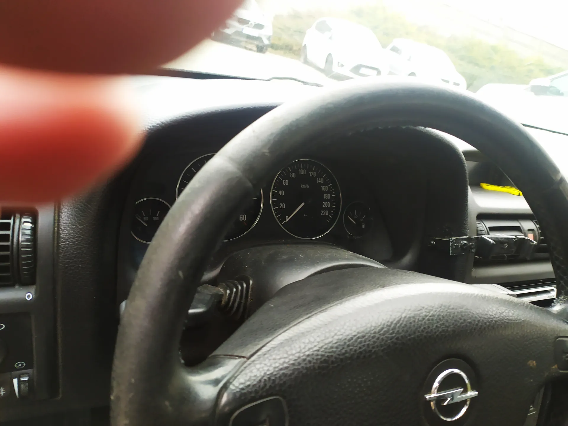Wnętrze samochodu z widocznym kierownicą marki Opel, tablicą przyrządów z prędkościomierzem (0-220 km/h), obrotomierzem i innymi wskaźnikami. W lewym górnym rogu widoczny fragment palca. Za przednią szybą parkujące samochody i zielone krzewy. Kierownica i tablica przyrządów są czarne, z widocznymi elementami deski rozdzielczej po lewej stronie.