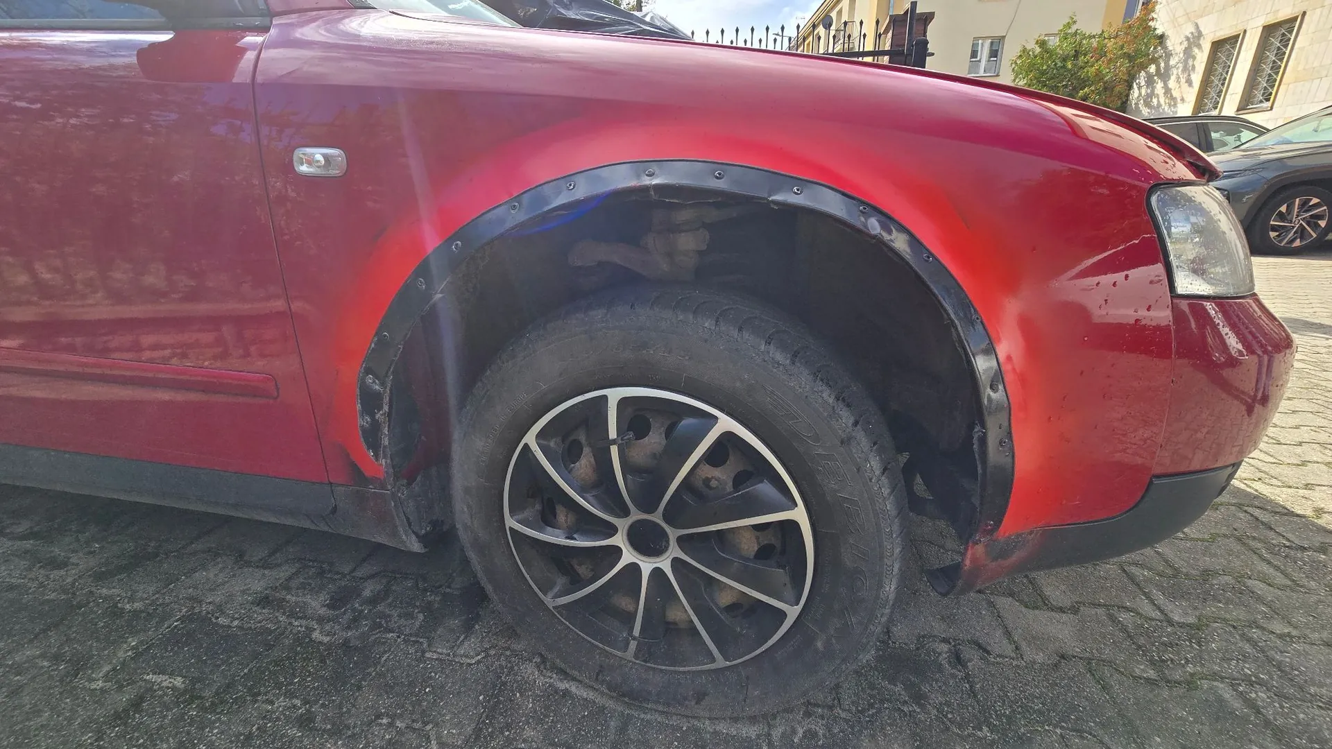 Zbliżenie na przednie prawe koło czerwonego samochodu. Nad oponą zamontowano czarny, plastikowy nadkole przykręcone śrubami do karoserii. Felga jest aluminiowa, wieloramienna, w kolorze srebrno-czarnym. W tle widoczny fragment chodnika z kostki brukowej oraz fragment budynku i innego samochodu.