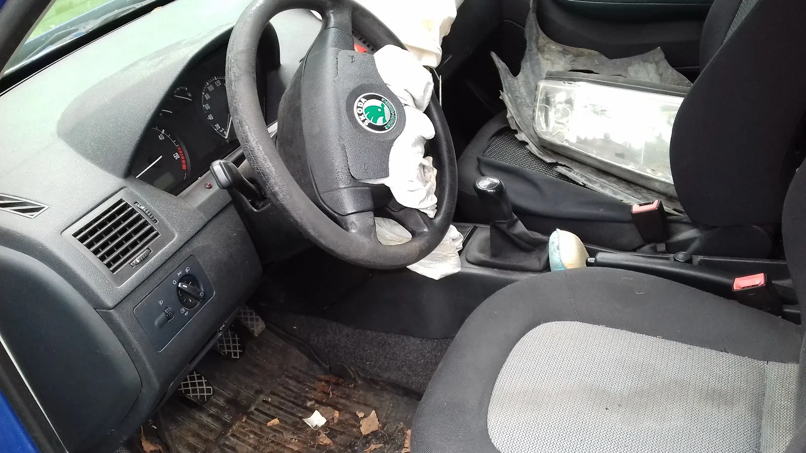 Wnętrze samochodu po wypadku. Widoczna jest deska rozdzielcza, kierownica z wystrzeloną poduszką powietrzną oraz dwa przednie fotele. W obszarze pasażera widoczne są znaczne zniszczenia, w tym rozbita przednia szyba i elementy wykończenia wnętrza. Na podłodze znajdują się zanieczyszczenia i drobne odłamki.