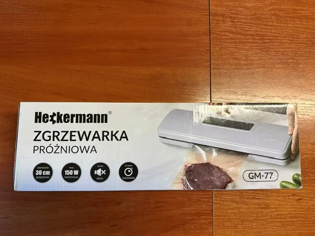 Opakowanie zgrzewarki próżniowej marki Heckermann, model GM-77. Na białym tle widnieje napis 'ZGRZEWARKA PRÓŻNIOWA' oraz zdjęcie szarego urządzenia do pakowania próżniowego z kawałkiem mięsa w folii. Poniżej znajdują się ikony z parametrami: szerokość zgrzewania 30 cm, moc maksymalna 150 W, cicha praca oraz szybkie zgrzewanie.