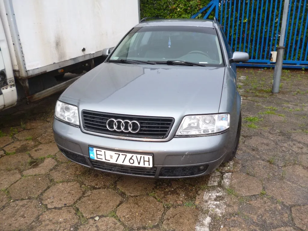 Srebrny samochód marki Audi zaparkowany na brukowanym podjeździe. Widoczny jest przód pojazdu z charakterystycznym grillem z czterema pierścieniami i polską tablicą rejestracyjną o numerze EL 776VH. Po lewej stronie znajduje się biała naczepa ciężarówki, a w tle niebieski metalowy płot i zielone krzewy.