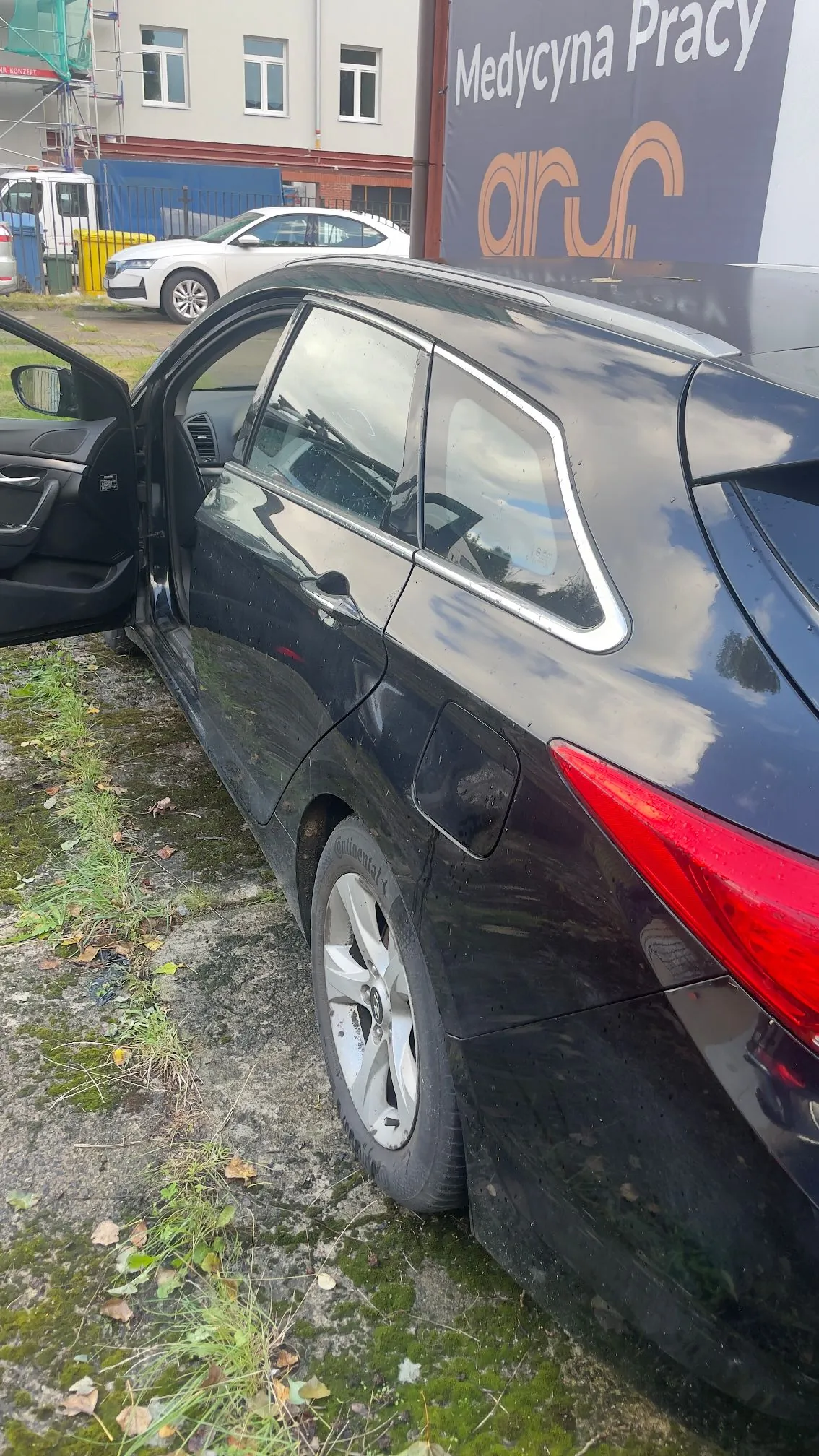 Czarny samochód typu kombi zaparkowany na trawiasto-betonowym podłożu. Przednie lewe drzwi są otwarte. W tle widoczny jest biały samochód, budynek z oknami oraz duży baner z napisem 'Medycyna Pracy' i logo 'aru'.