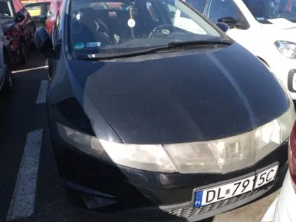 Czarny samochód marki Honda zaparkowany na miejscu parkingowym. Widoczna tablica rejestracyjna PL DL 791 SC, reflektory, przednia szyba z elementami wewnątrz oraz fragmenty innych pojazdów po bokach. Asfalt z białymi liniami wyznaczającymi miejsca parkingowe.