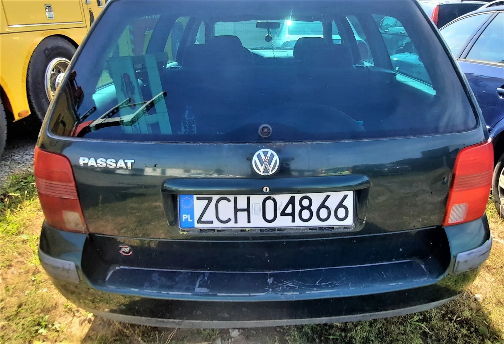 Tylny widok ciemnozielonego samochodu Volkswagen Passat. Na klapie bagażnika znajduje się napis 'PASSAT', logo Volkswagena oraz polska tablica rejestracyjna o numerze 'ZCH 04866'. Po lewej stronie widoczny jest fragment żółtego pojazdu.