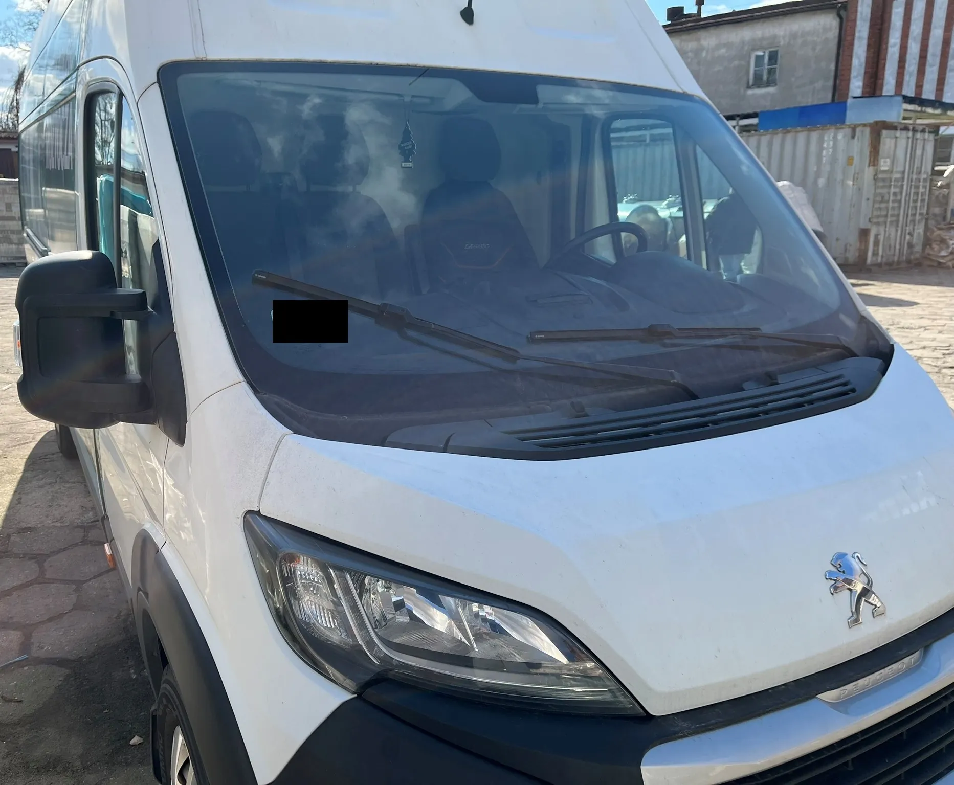 Zbliżenie na przód białego vana marki Peugeot. Widoczny jest przedni reflektor, logo lwa na masce oraz przednia szyba z wycieraczkami. Wewnątrz pojazdu widoczne są fotele i kierownica. Część szyby jest zakryta czarnym prostokątem. W tle znajduje się budynek i ogrodzenie.