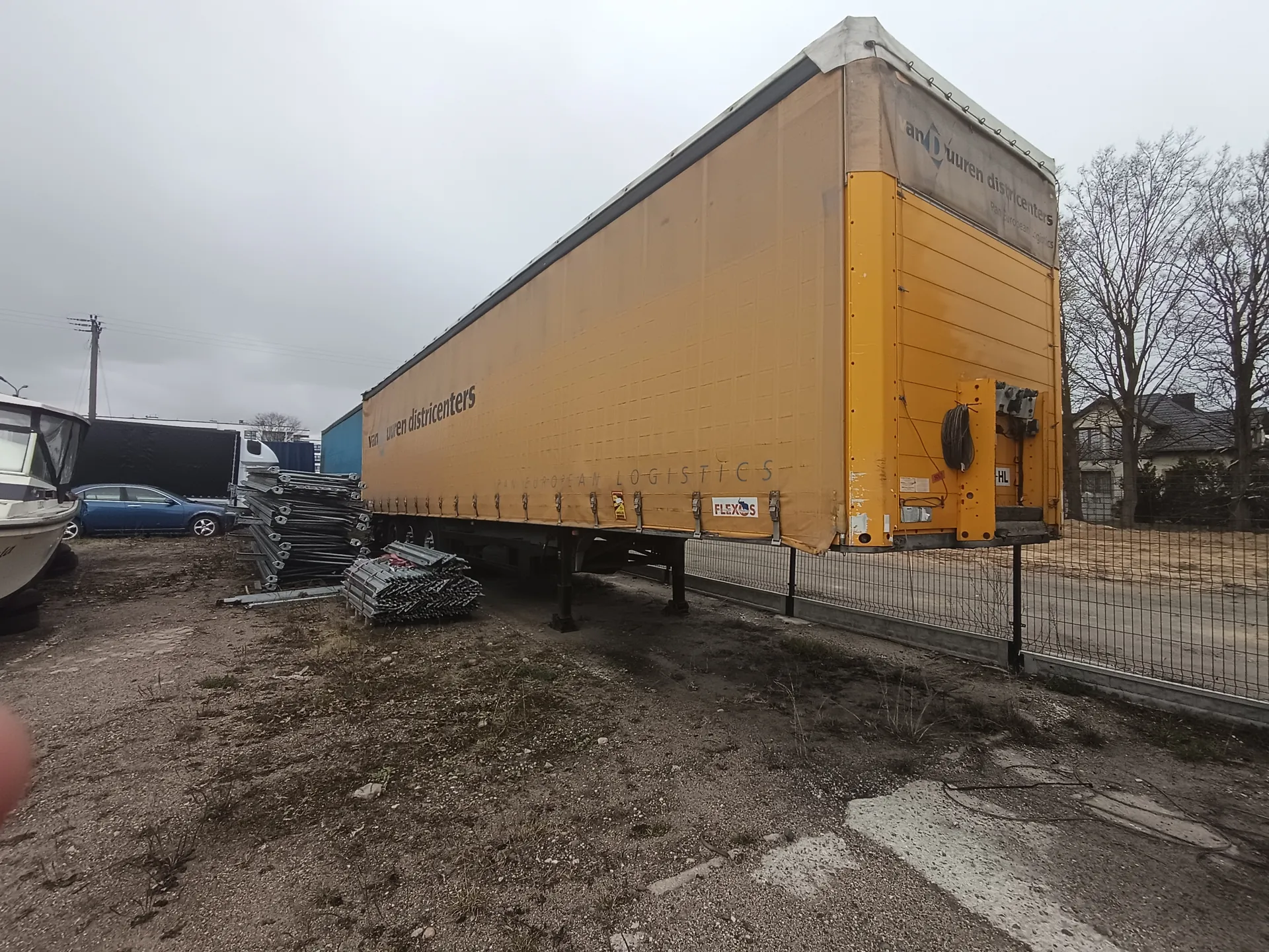 Duży żółty naczepa ciężarowa z napisami 'van uuren distributiecenters', 'EUROPEAN LOGISTICS' oraz logo 'FLEXOS'. Na tylnej części naczepy widoczne są dodatkowe napisy 'and uuren distributiecenters' i oznaczenie 'HL'. Naczepa stoi na podwórku z żwirem, obok metalowego ogrodzenia. W tle: niebieski samochód, łódź, stosy metalowych ramek, drzewa bez liści, domy i pochmurne niebo.