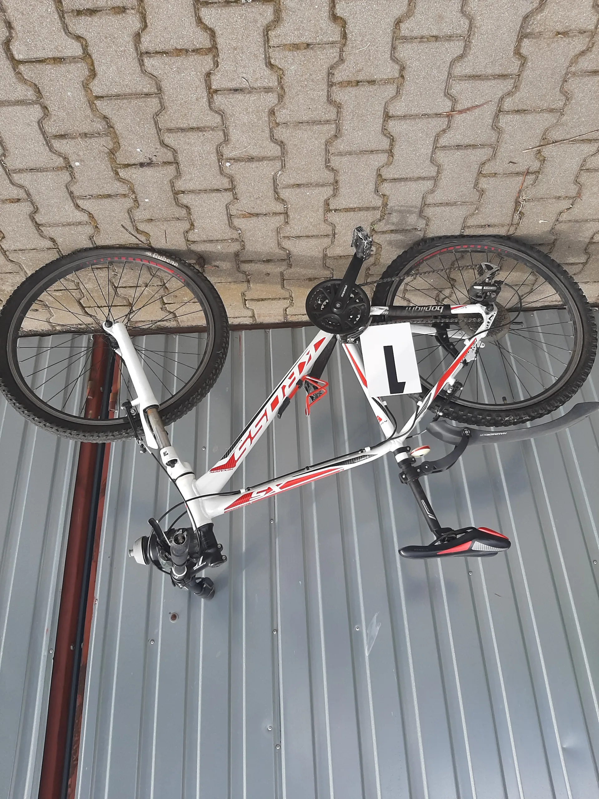 Biały rower z czerwonymi akcentami oparty o metalową ścianę z pionowymi listwami i brukowaną powierzchnię. Na ramie widoczny napis 'KROSS', a na białej tabliczce czarna cyfra '1'. Rower jest ustawiony pionowo, z kołami w pozycji pionowej. Siedzisko jest czarno-czerwone, a na kierownicy znajduje się dzwonek.