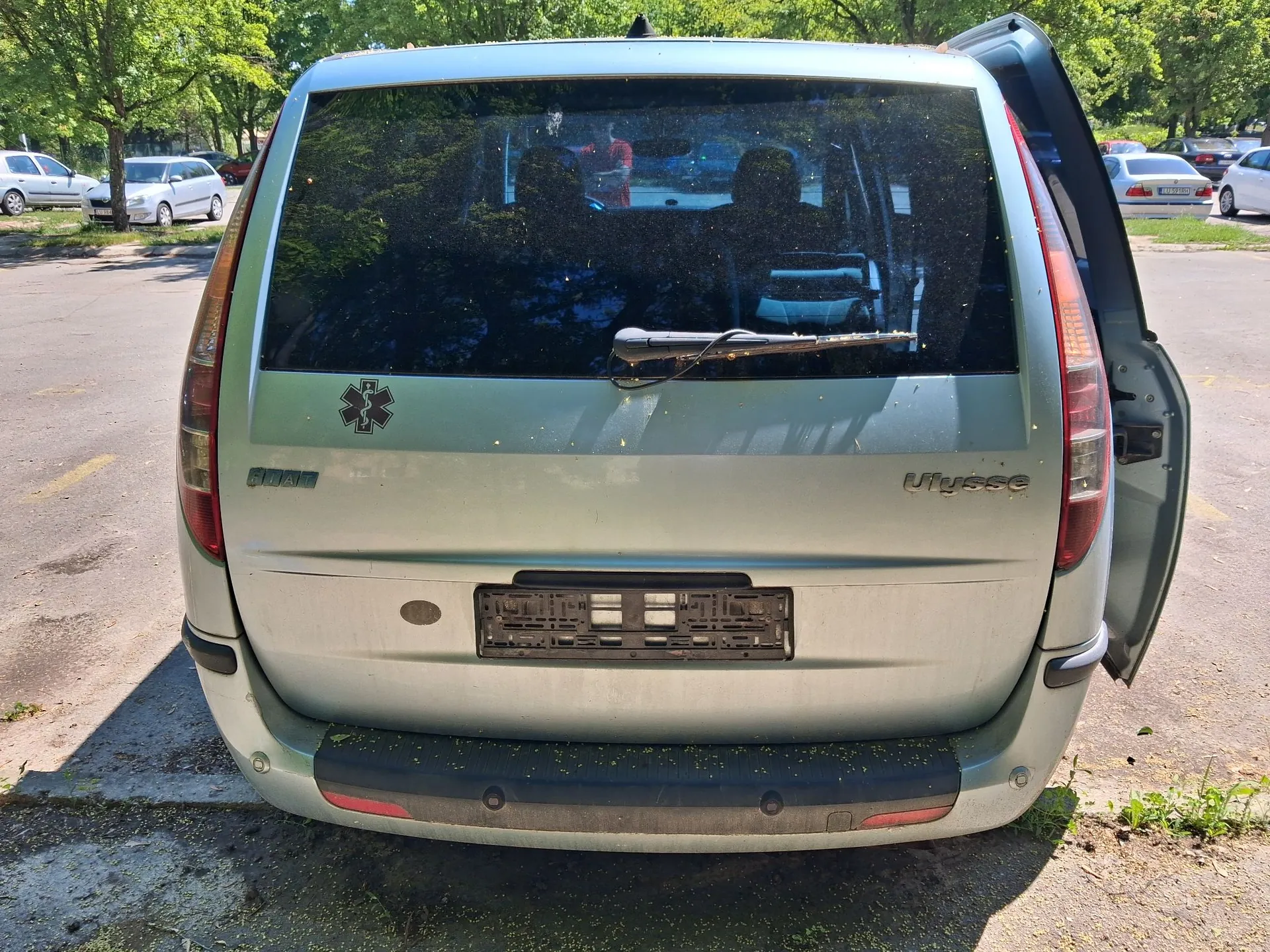 Tylny widok srebrno-zielonego samochodu typu minivan z otwartą klapą bagażnika. Na klapie znajduje się napis 'Ulysse', logo marki oraz naklejka z symbolem gwiazdy życia (Star of Life). Samochód nie posiada tablic rejestracyjnych, a w miejscu ich montażu widoczna jest czarna ramka. Pojazd stoi na parkingowym placu w otoczeniu drzew.