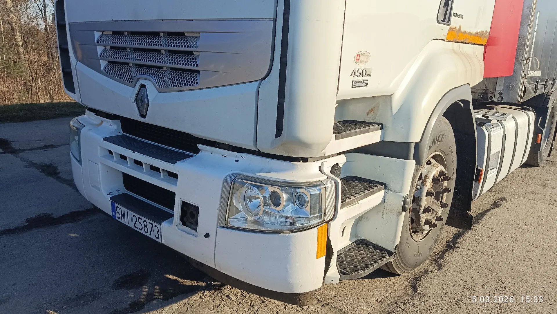 Zbliżenie na przód białej ciężarówki marki Renault. Widoczny jest fragment białego zderzaka z tablicą rejestracyjną o numerze SMI 2587-3, reflektor oraz grill z logo producenta. Na boku kabiny znajduje się napis '450 m.hp EURO 5'. Pojazd stoi na utwardzonej nawierzchni.
