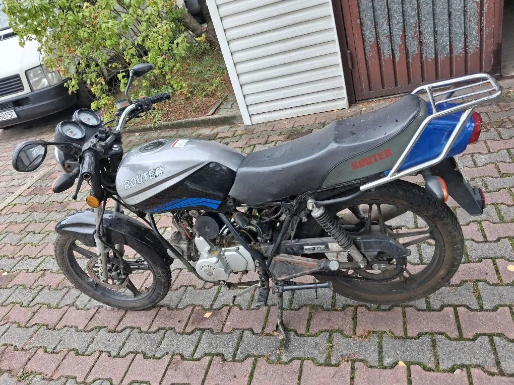 Srebrno-niebieski motocykl marki Router zaparkowany na kostce brukowej. Pojazd posiada czarne siedzenie, tylny bagażnik oraz widoczny silnik. W tle znajduje się biała roleta garażowa, brązowe drzwi oraz fragment białego samochodu i zielone krzewy.