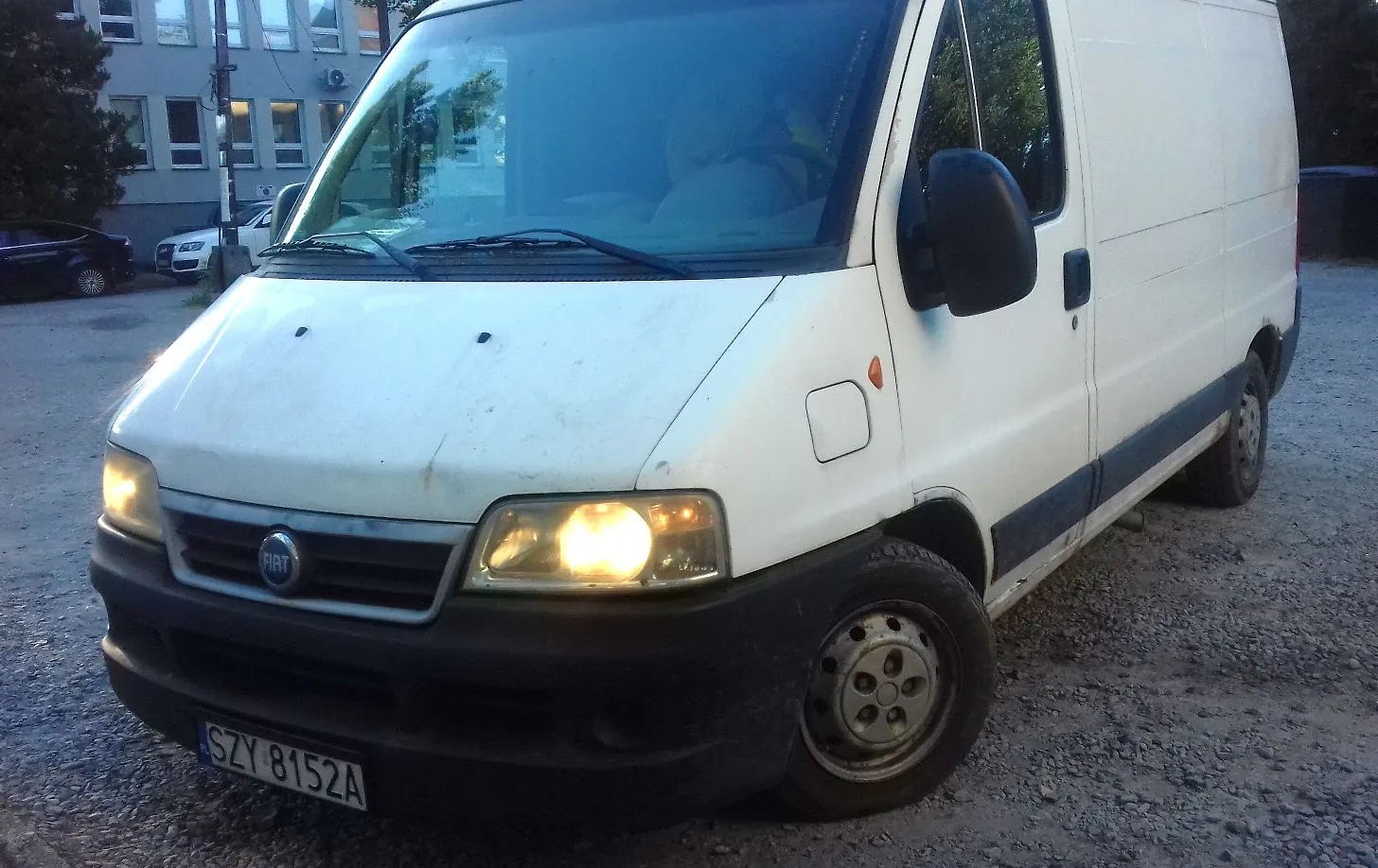 Biały furgon marki Fiat zaparkowany na żwirowym podłożu. Pojazd jest widoczny z przodu i z boku, z włączonym prawym reflektorem. Na przednim zderzaku znajduje się tablica rejestracyjna o numerze SZY 8152A. W tle widoczny jest budynek oraz inny samochód.