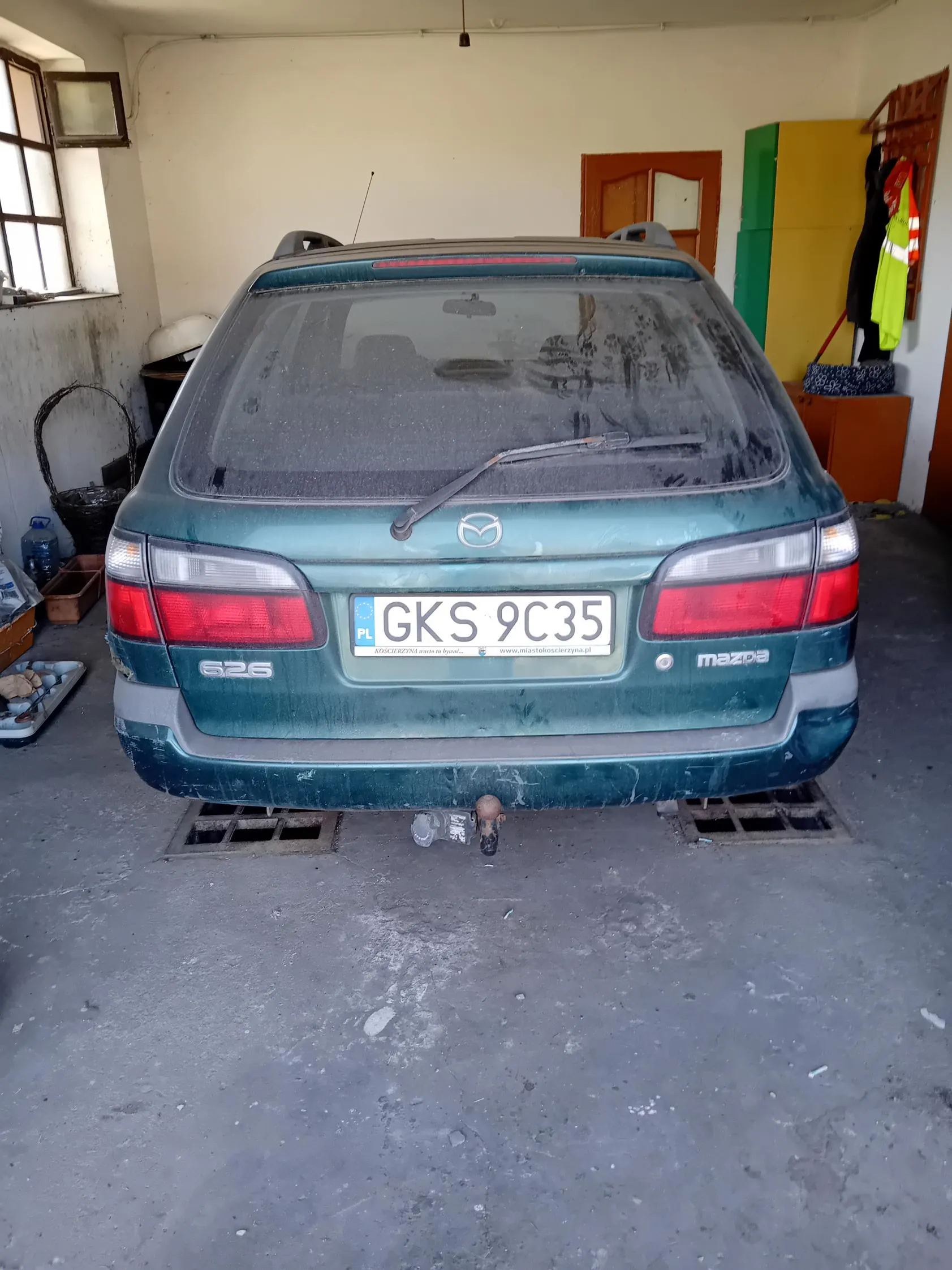 Z tyłu zielony samochód Mazda 626 z tablicą rejestracyjną PL GKS 9C35. Na tablicy widoczny napis www.miastokoscierzyna.pl. Samochód stoi w garażu z betonową podłogą, z lewej strony okno, kosz, pojemnik z wodą i narzędzia. Z prawej strony kolorowa szafka (zielona i żółta), drzwi oraz wiszące kamizelki odblaskowe. Tylne szyby samochodu są zabrudzone, widoczny wycieraczka i hak holowniczy. Na bagażniku napisy '626' i 'mazda'.