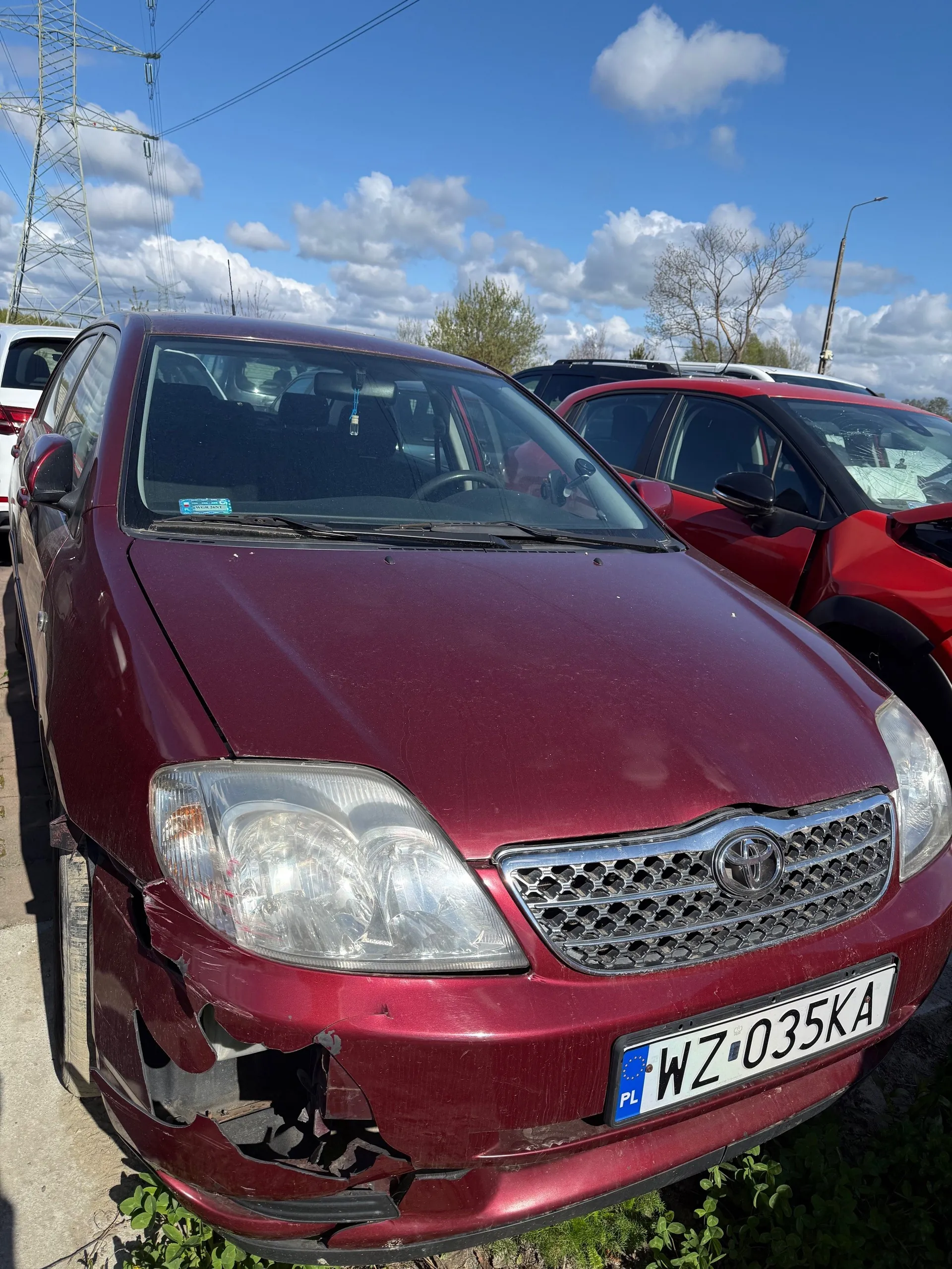Uszkodzony bordowy samochód marki Toyota z tablicą rejestracyjną WZ 035KA (PL) na przodzie. Widoczne uszkodzenie pasa przedniego, w tym zdeformowany zderzak i reflektory. W tle inne pojazdy, w tym czerwony samochód, niebo z chmurami oraz maszt energetyczny. Na przedniej szybie niebieska naklejka.