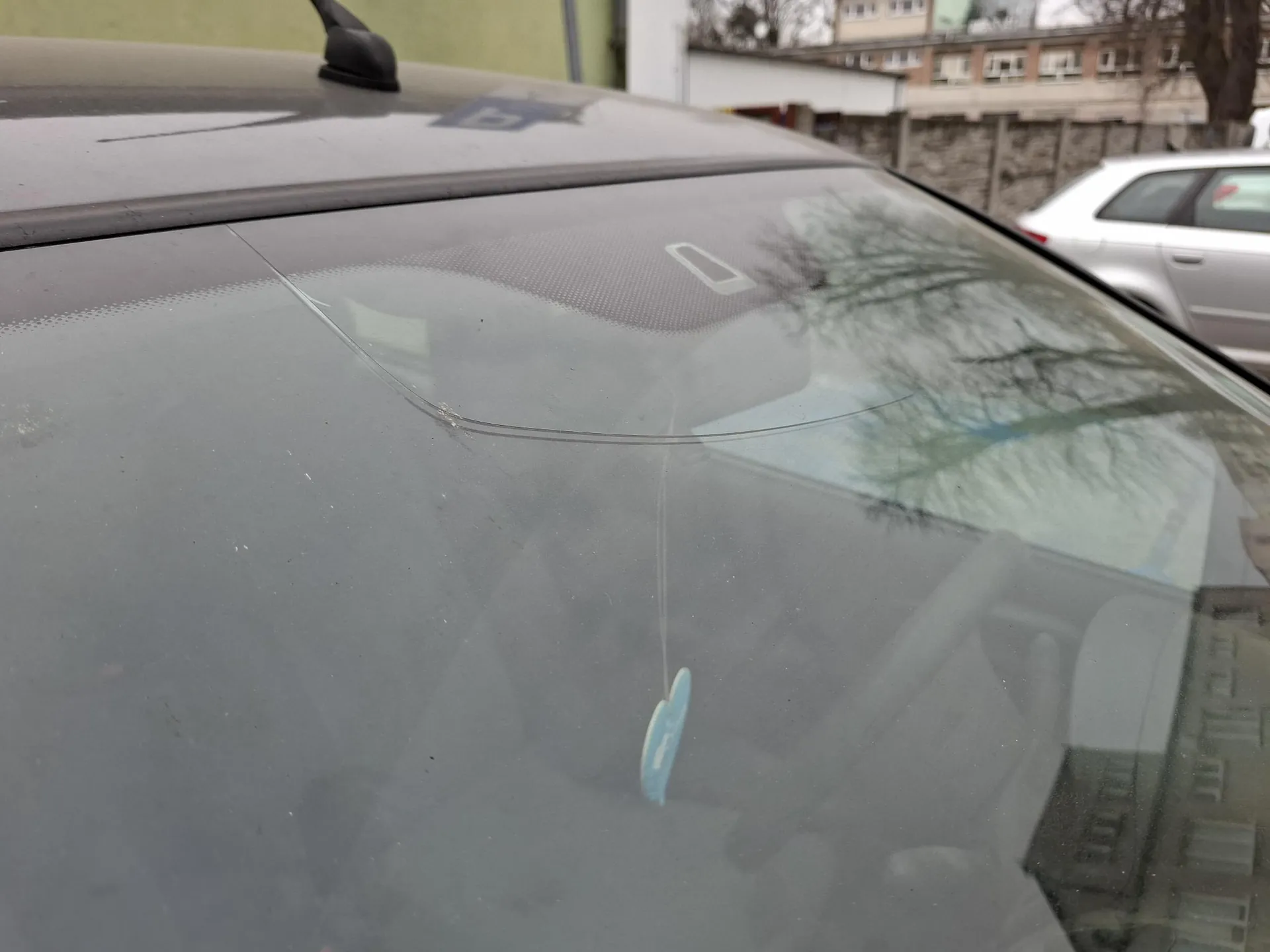 Zbliżenie na przednią szybę szarego samochodu z widoczną pionową pęknięciem. Na szybie wiszący niebieski element ozdobny lub znaczek. W tle częściowo widoczny srebrny samochód, kamienica z oknami oraz drzewa bez liści. Na dachu szarego samochodu widoczny czarny akcesorium.