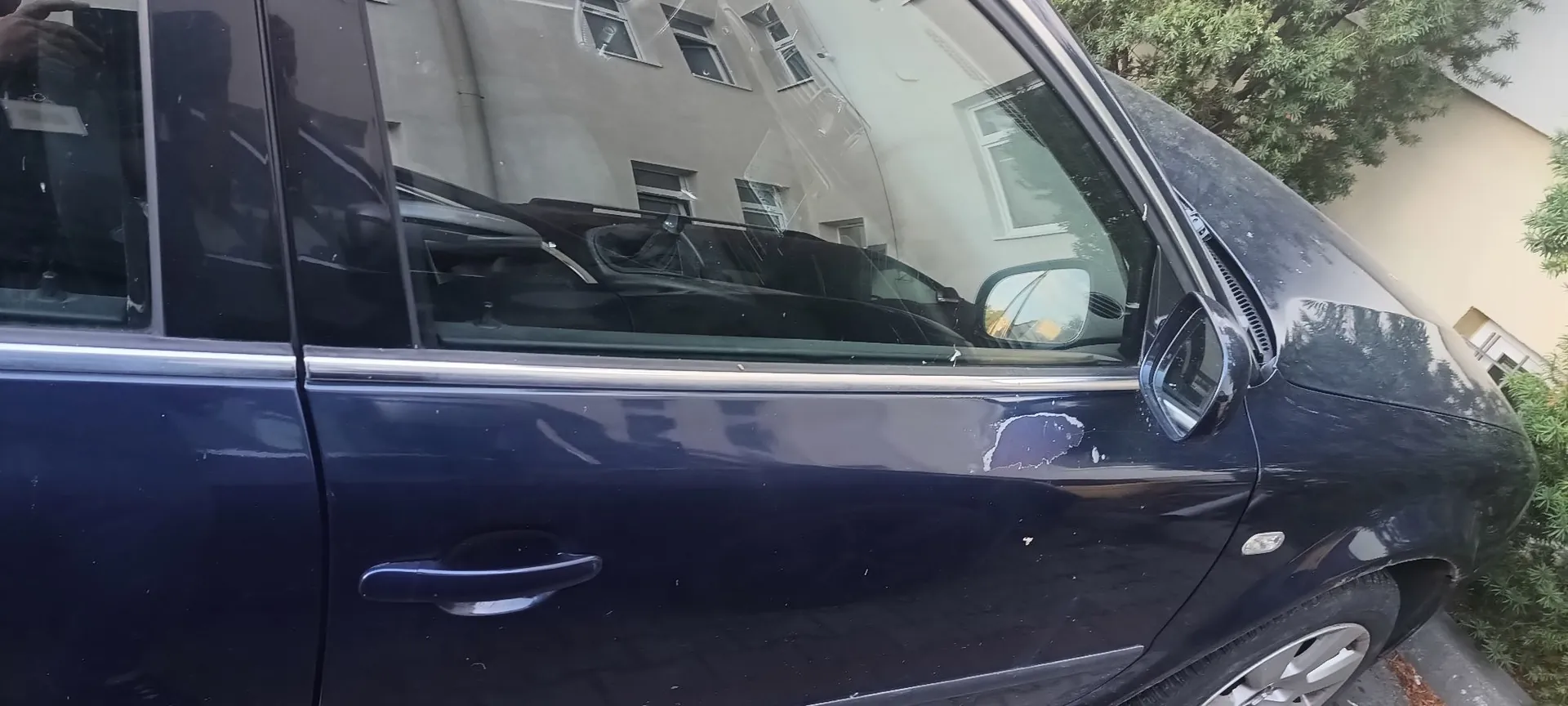 Zbliżenie na ciemnoniebieski samochód. Na przednich drzwiach kierowcy widoczne jest uszkodzenie lakieru w postaci jasnej zadrapania lub odprysku. W odbiciu szyb samochodu widać jasny budynek mieszkalny oraz drzewa.