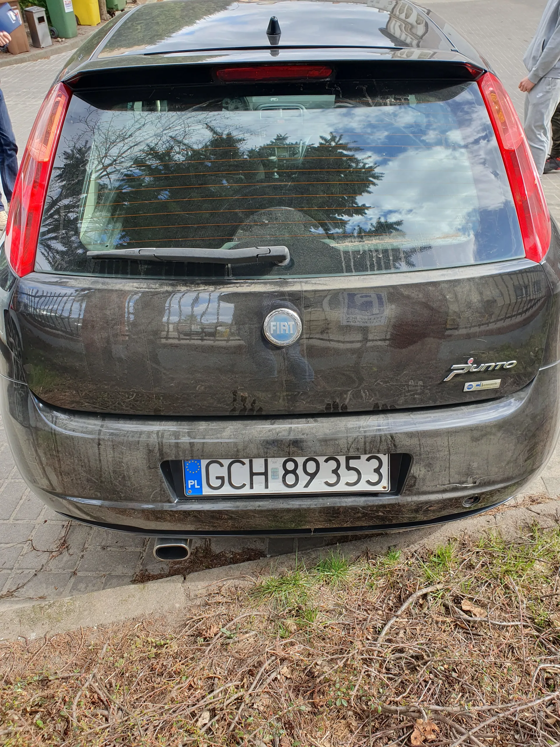Tył ciemnego samochodu marki Fiat model Punto z tablicą rejestracyjną GCH 89353 PL. Samochód zaparkowany na chodniku obok suchych krzewów i trawy. W tle odbicia drzew i nieba w szybie. W lewym górnym rogu fragment ręki osoby.