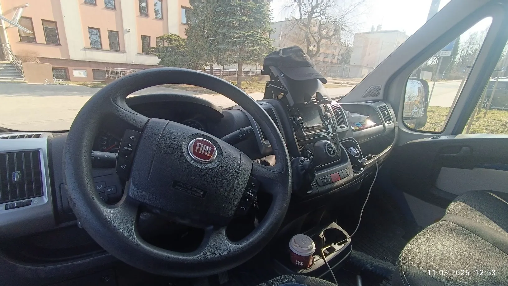 Wnętrze samochodu marki Fiat widziane z perspektywy kierowcy. W centrum znajduje się czarna kierownica z logo FIAT na środku. Widoczna jest deska rozdzielcza z radiem, przyciskami sterującymi oraz uchwytem na telefon zamontowanym na górze. W uchwycie znajduje się ciemny przedmiot. W pod Cup holderze widać kubek z kawą. Przez przednią szybę widoczny jest budynek mieszkalny oraz drzewa. W prawym dolnym rogu znajduje się data i godzina: 11.03.2026 12:53.