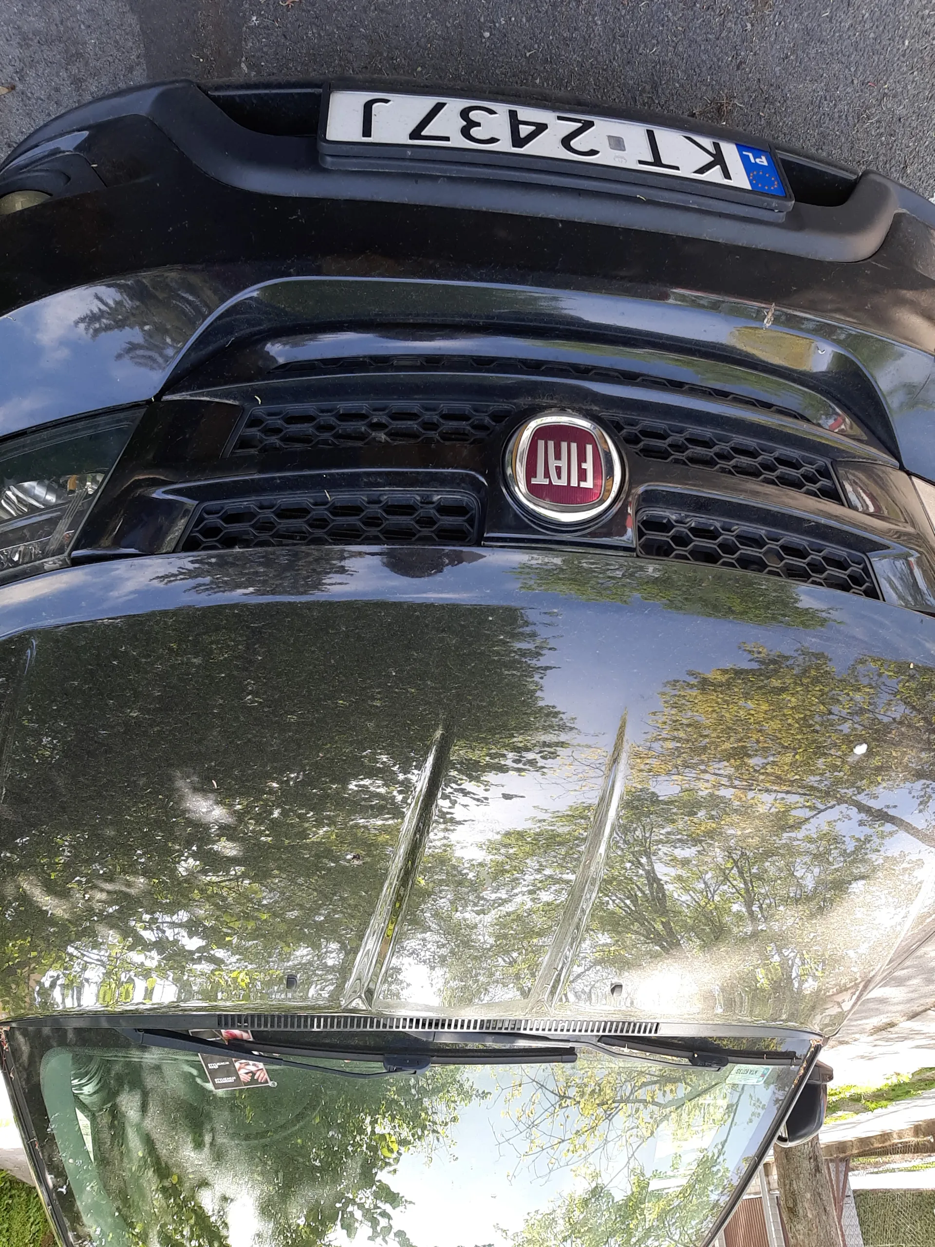 Czarny samochód marki Fiat z czerwonym logo na atrapie chłodnicy. Tablica rejestracyjna z białym tłem i czarnym tekstem 'KT 2437 J' oraz niebieskim pasem z literą 'PL' po lewej stronie. Na przedniej szybie widoczne są wycieraczki i odbicie drzew oraz nieba. Samochód zaparkowany na asfaltowej powierzchni.