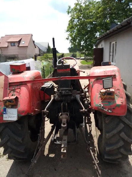 Widok z tyłu na stary, czerwony traktor rolniczy zaparkowany na ziemnym podłożu. Maszyna posiada duże, błotniste opony z charakterystycznym bieżnikiem. W tle widoczny jest dom z brązowym dachem, biały budynek oraz zielone drzewa pod jasnym niebem.